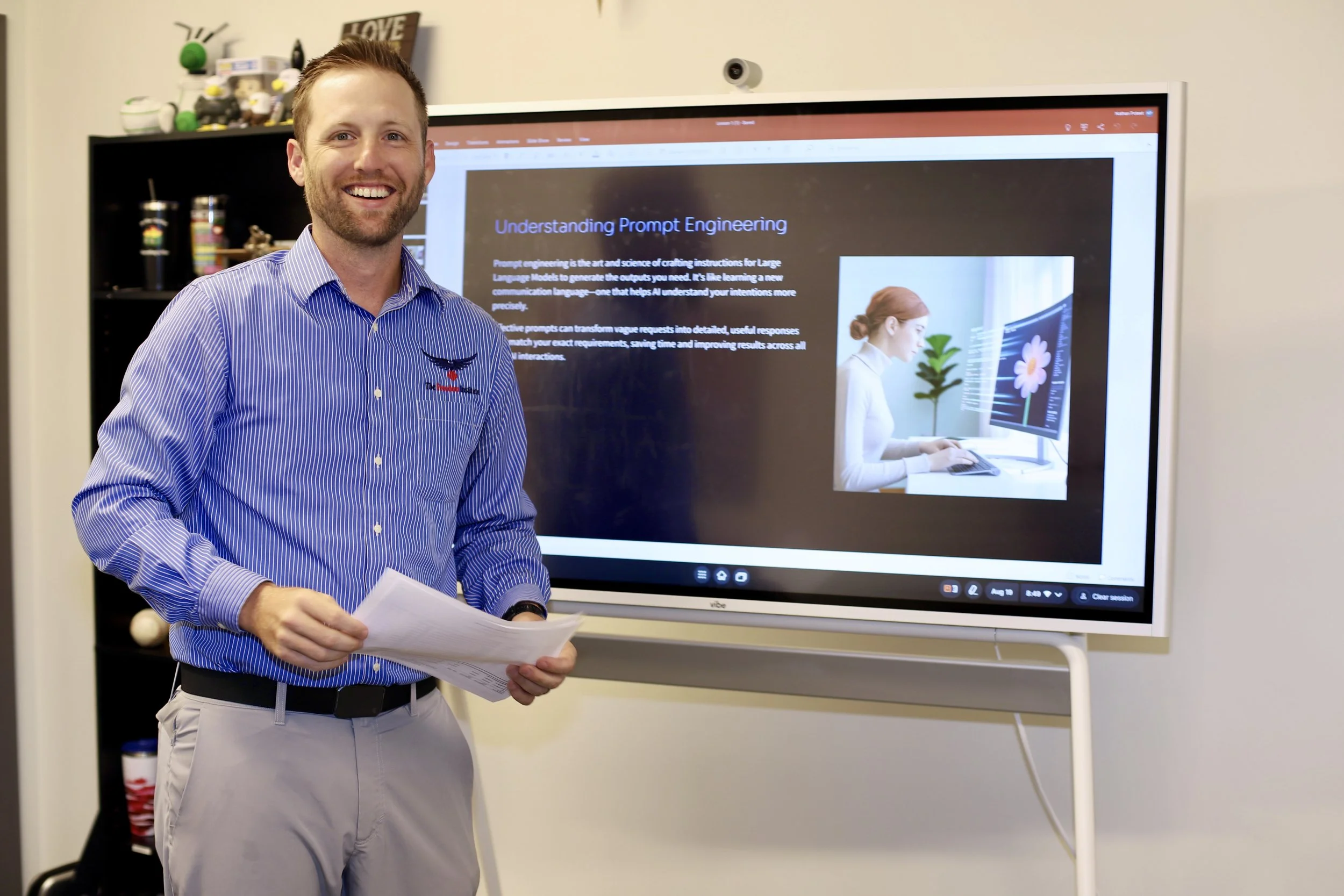 A man in a blue-striped shirt presenting in front of a digital display screen with a presentation slide about prompt engineering, holding papers in his hand and smiling.