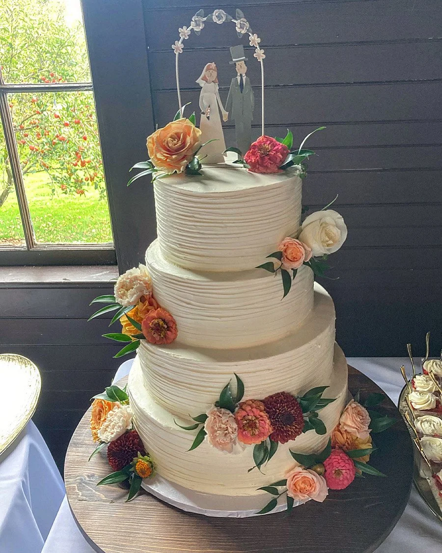 A three-tier white wedding cake decorated with pink, red, and cream-colored flowers and green leaves. A cake topper features a paper cutout of a bride and groom holding hands under an arch of flowers.