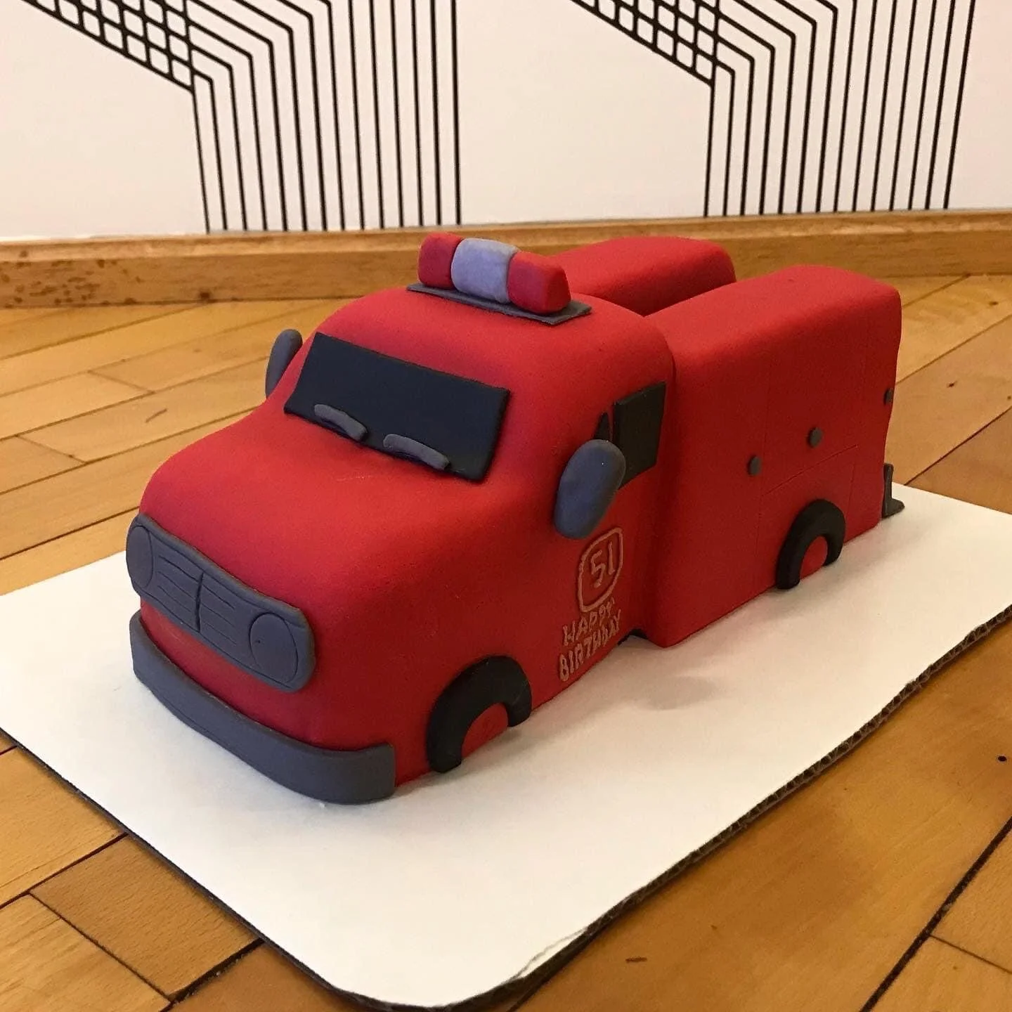 Cake shaped like a red fire truck with black and gray details, placed on a white board on a wooden table.