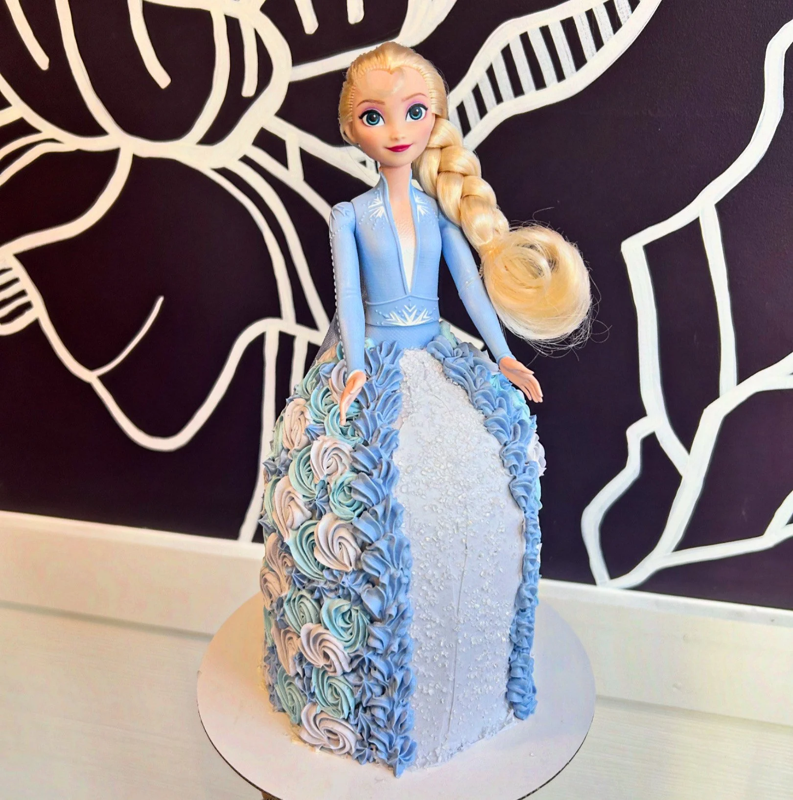 A cake designed to look like Elsa from Disney's Frozen, with a figurine of Elsa on top. The cake features blue, white, and pastel-colored roses on the skirt and has a black and white floral background.