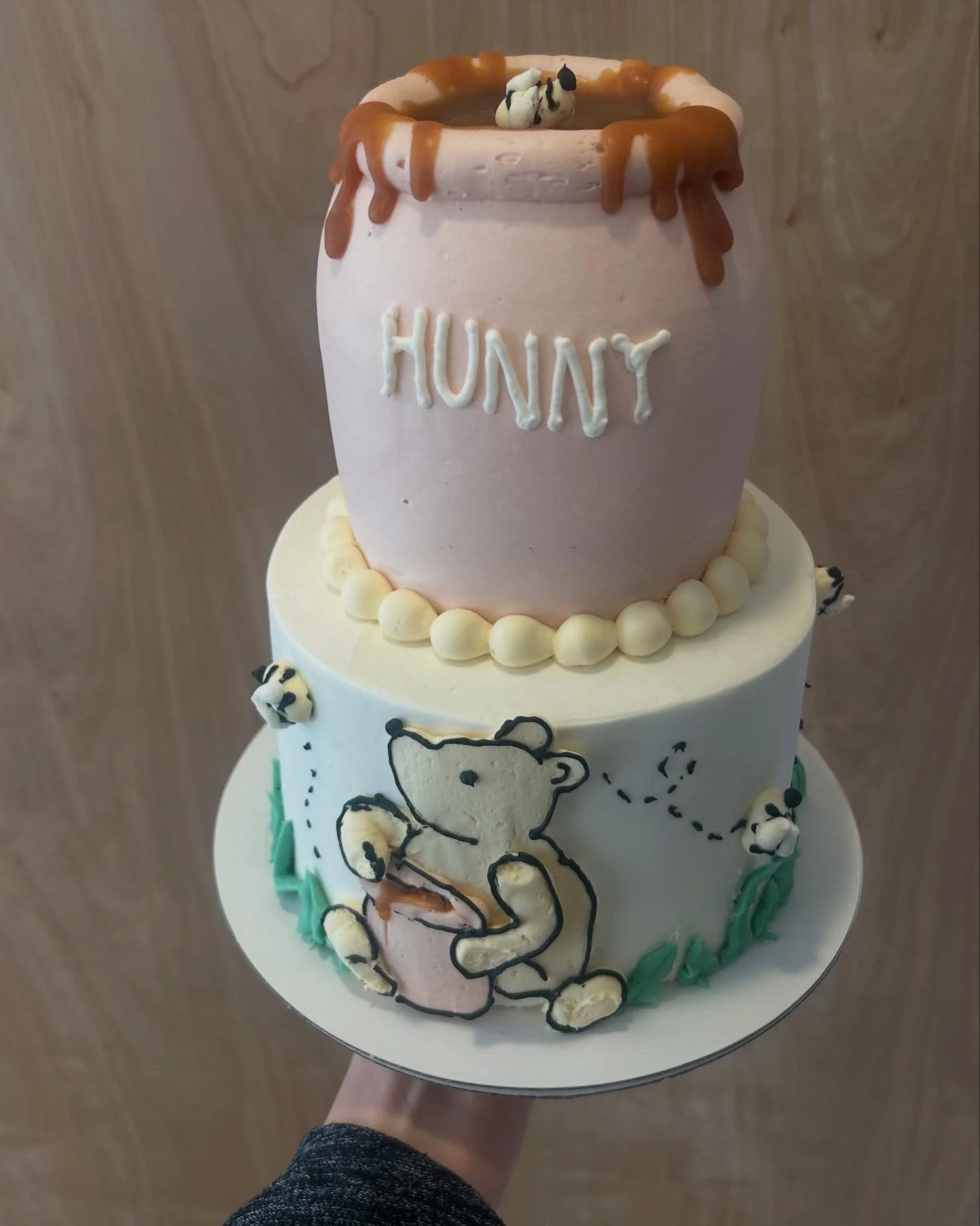 A two-tiered birthday cake with a bear holding a basket and the word 'HUNNY' on the top tier, decorated with a pink and white color theme, bear and honeycomb decorations, and green icing at the bottom.