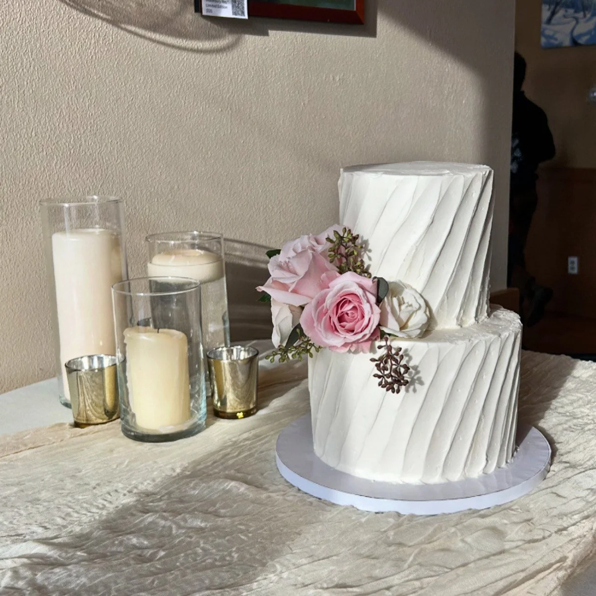 Two-tier white wedding cake with pink and white roses and greenery, surrounded by four candles in glass holders and a beige table runner.