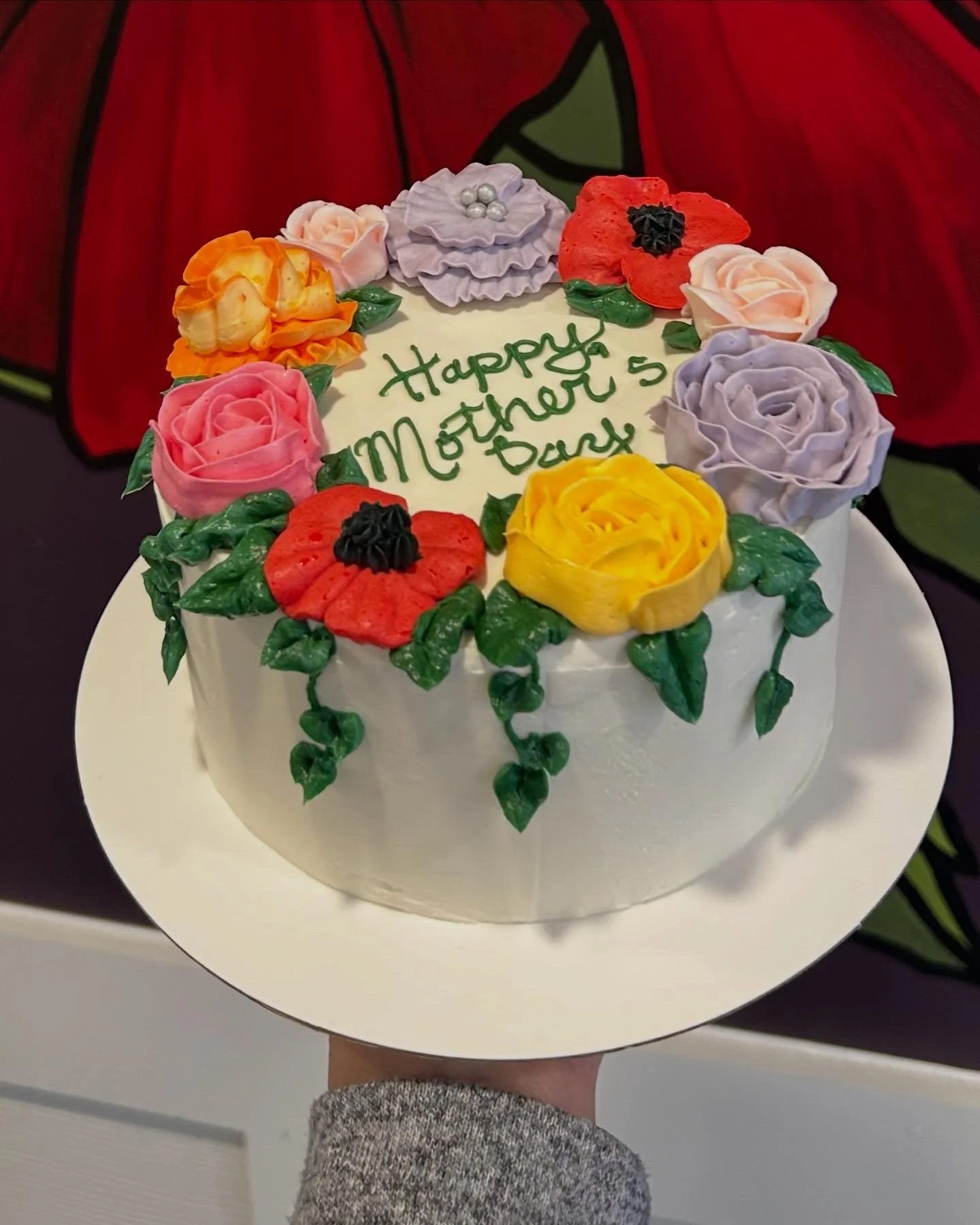 A white frosted cake decorated with colorful fondant flowers and green leaves. The cake has a message written in green icing that says, 'Happy Mother's Day 5.' The background features a red and green design.