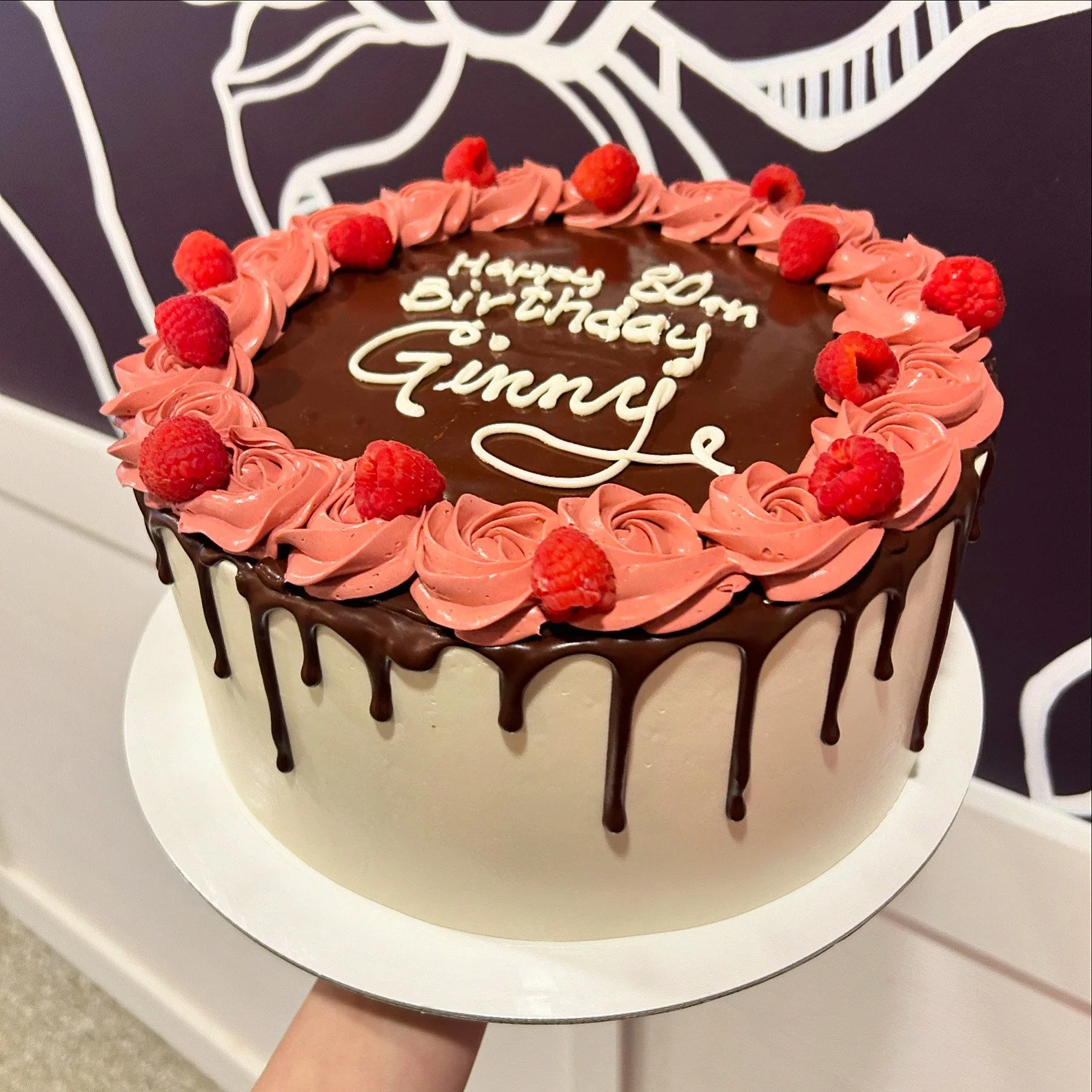 A round birthday cake decorated with pink frosting roses and raspberries, with a chocolate drip on the sides. The top of the cake has a message written in white icing that says, 'Happy 30th Birthday Gimmie'.