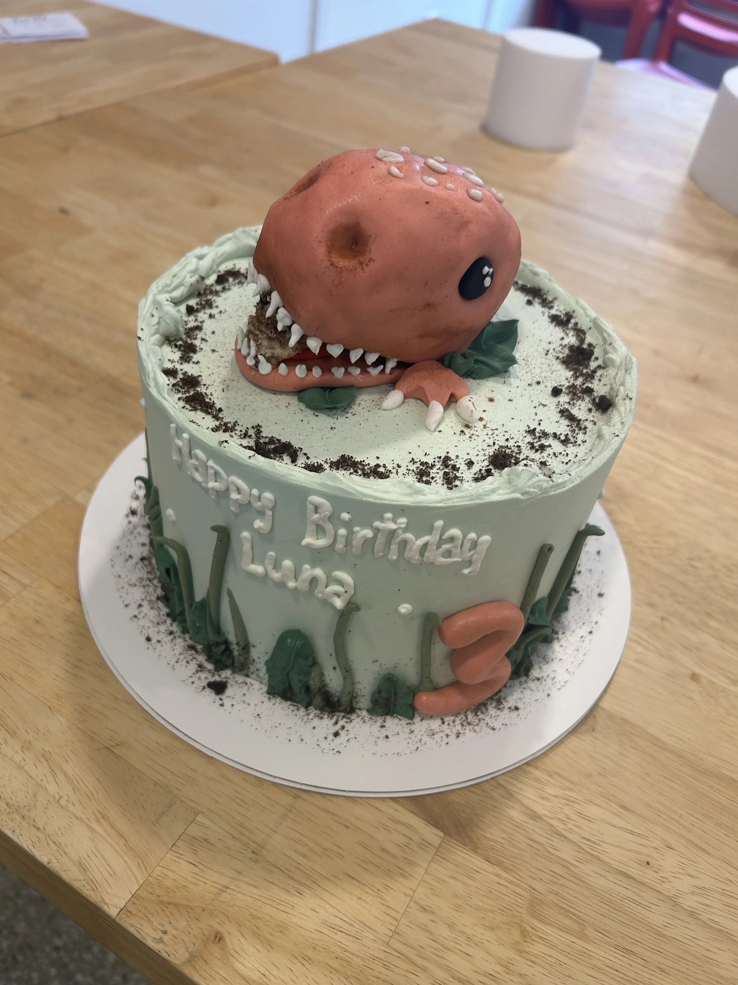 A dinosaur-shaped cake with light green frosting, decorated with white and dark green accents, and a large, pink dinosaur head with white teeth, black eye, and small white spots. The cake has the message 'Happy Birthday Luna' written on it and the nu