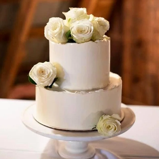 Two-tier white wedding cake decorated with white roses on top and at the side, placed on a white cake stand.
