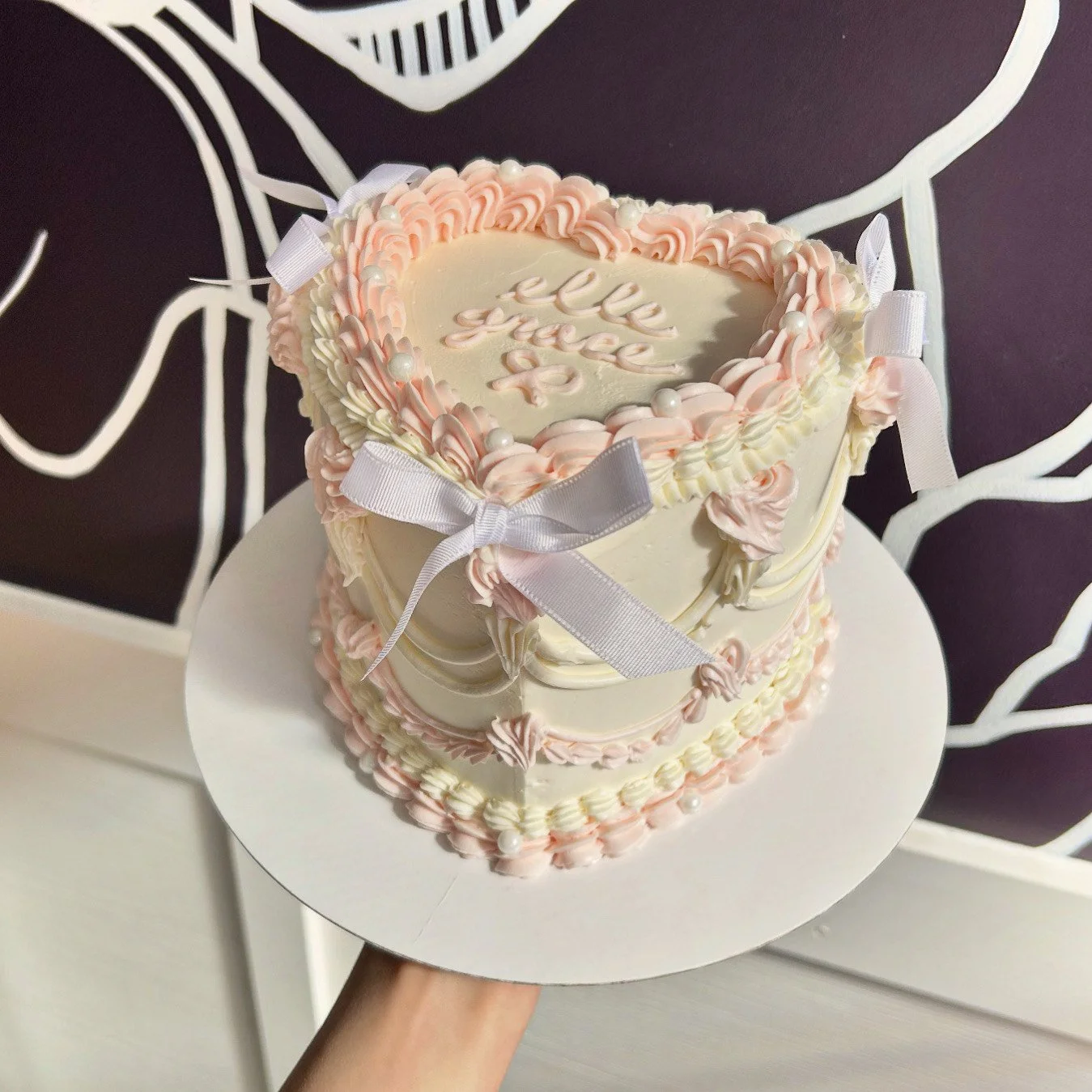 A heart-shaped cake decorated with pink and white frosting, pearls, and a white ribbon. The top has a message written in pink icing that says 'lee lee 30p'.