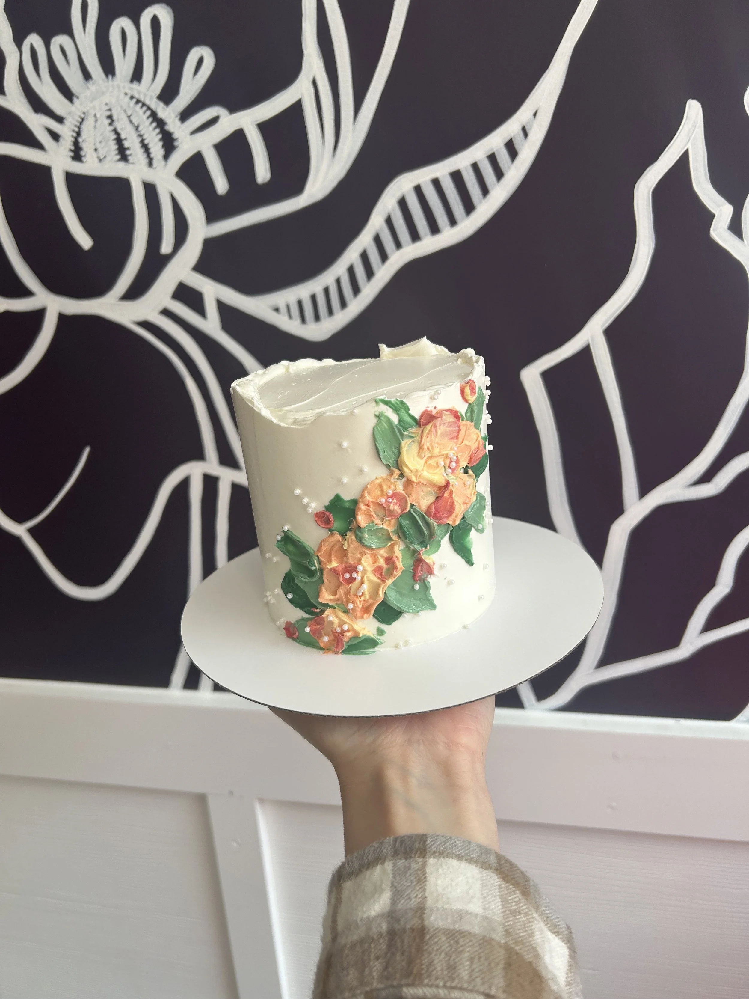 Cake with white frosting decorated with orange and yellow flowers and green leaves, held on a white cake board against a blackboard with white chalk flower illustrations.