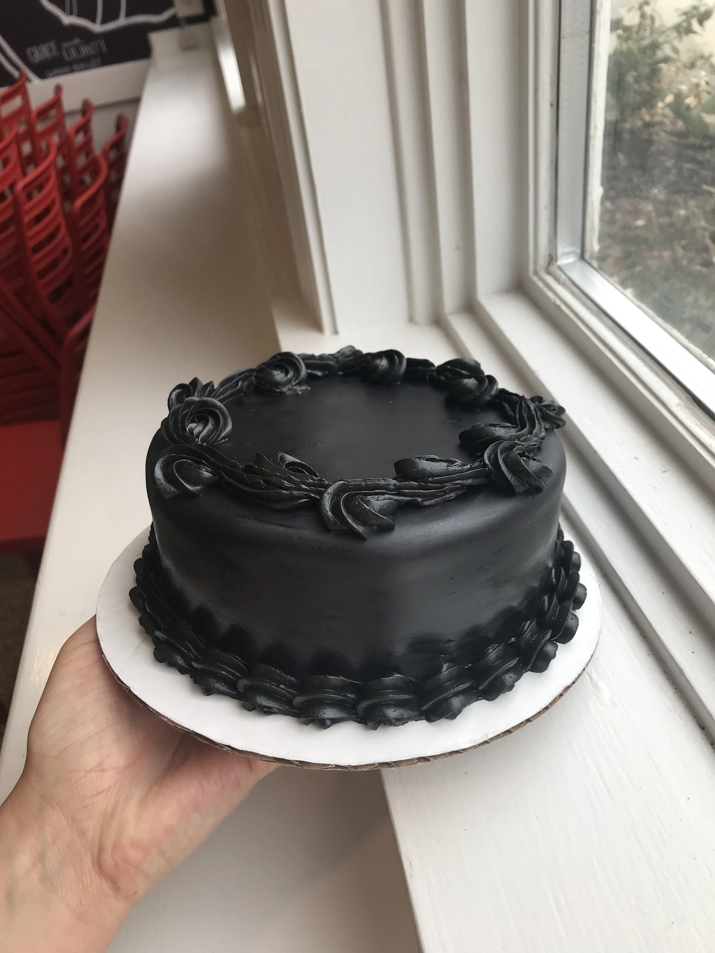 A black frosted cake with decorative piping on top and around the base, held near a window with white trim.