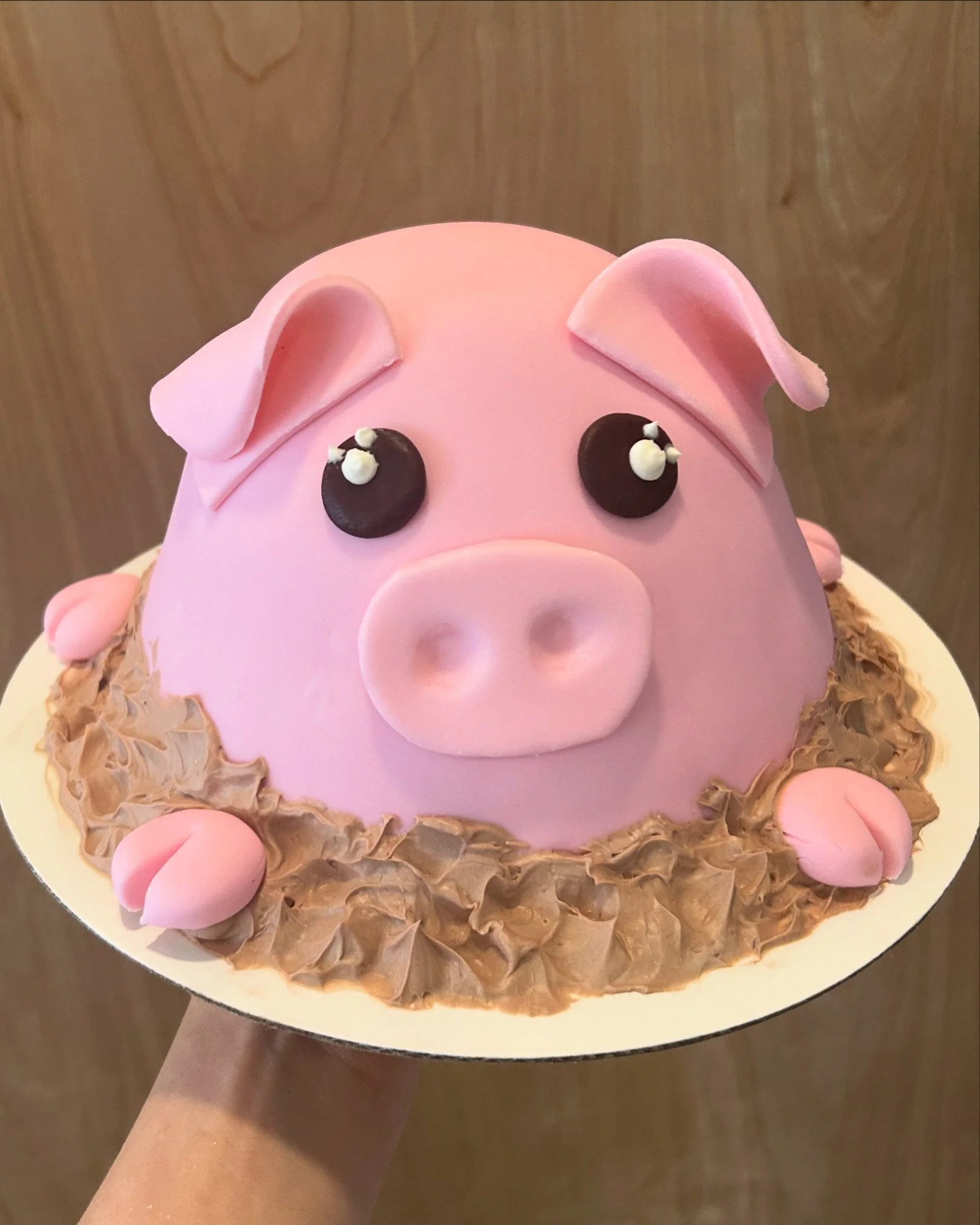 A pig-shaped cake with pink fondant, black candy eyes, pink fondant ears, a snout, and surrounded by brown and pink decorative elements.