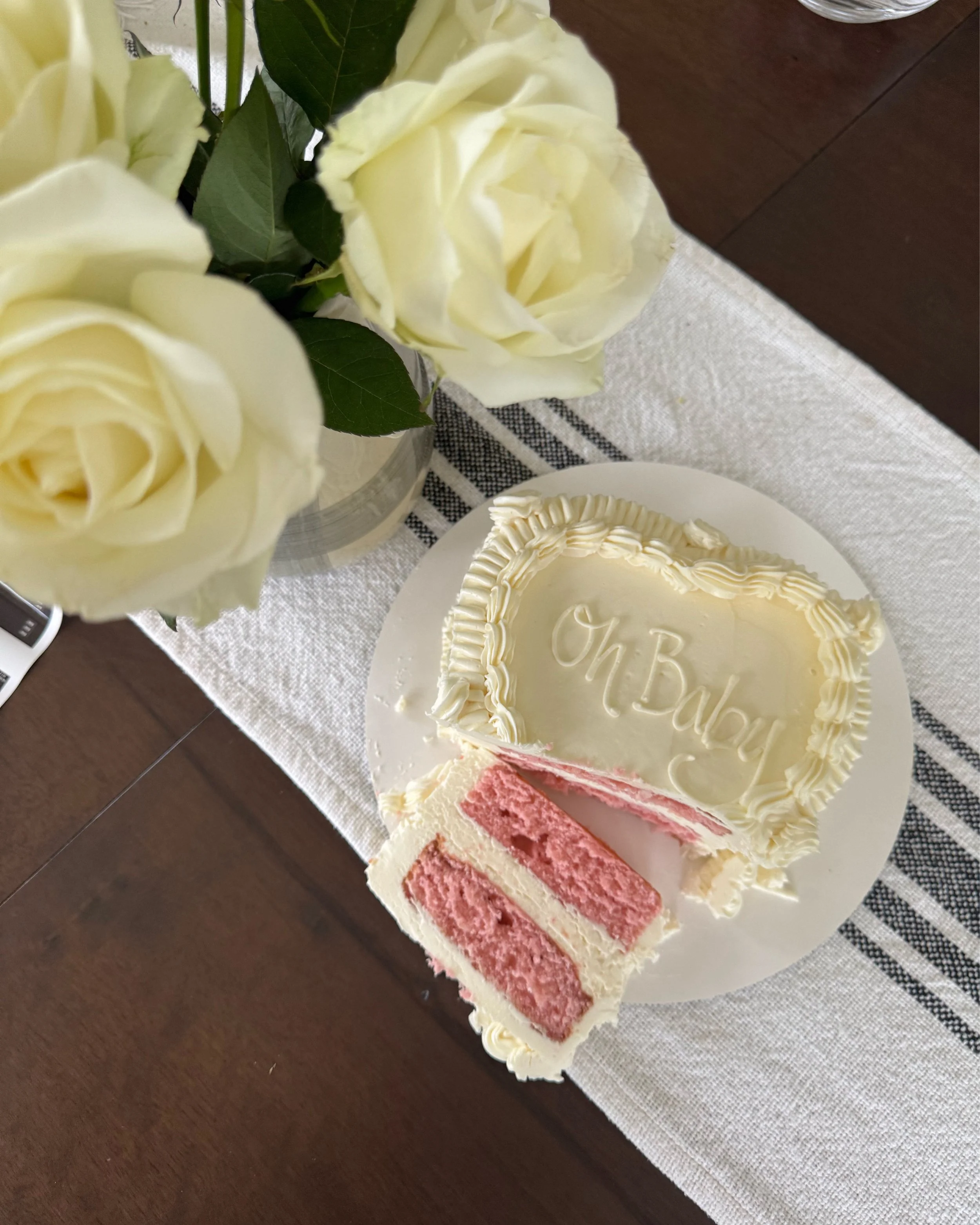 A birthday cake with white icing and the message "Oh Baby" written on top, along with a slice showing pink and white layers. A vase of white roses is placed nearby on a striped cloth.