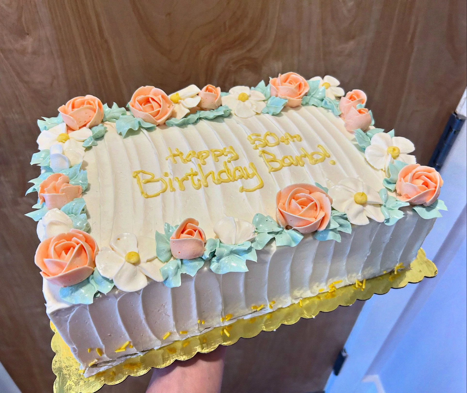 A rectangular birthday cake decorated with pastel pink and white flowers and green leaves, with yellow writing that says 'Happy 50th Birthday Barb!'