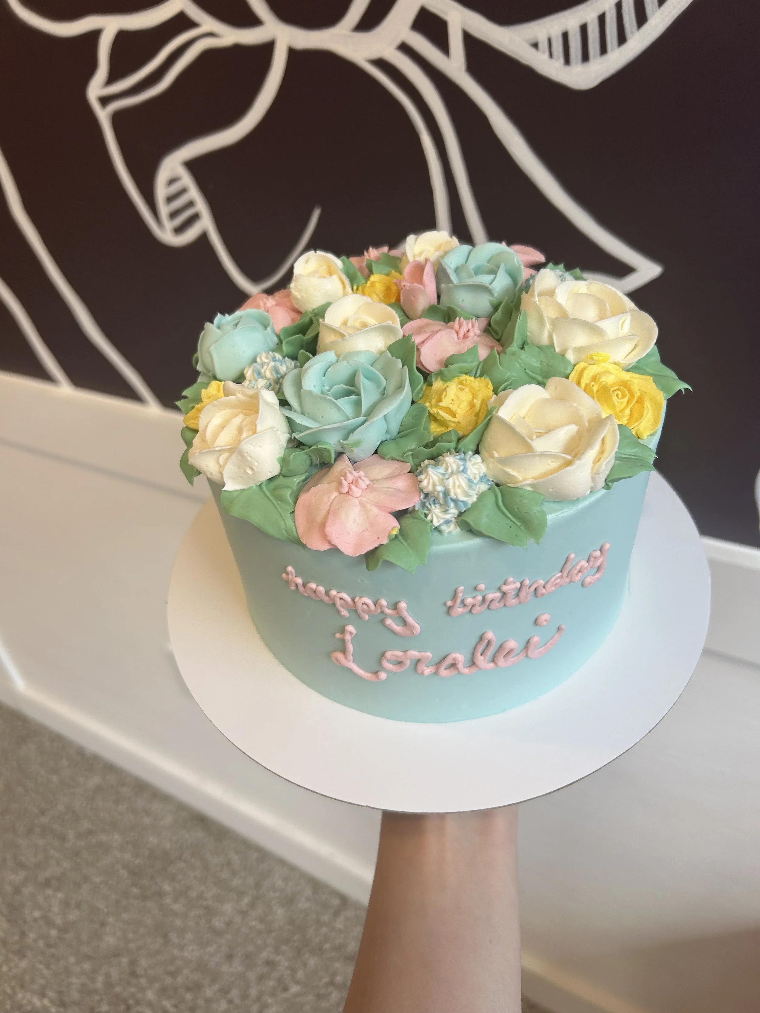 A round birthday cake decorated with pastel-colored flowers and green leaves. The cake has a message written in pink icing that reads "happy birthday Lorelei." The cake is held on a white cake board by a person, with a black and white wall art in the