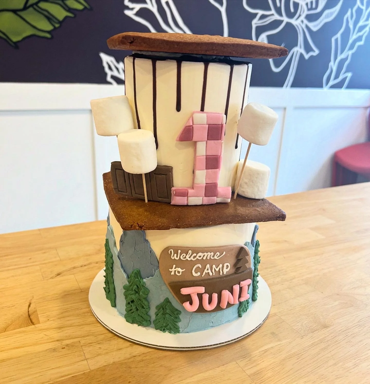 A camping-themed cake with a brown top and white sides decorated with green trees and a sign that says "Welcome to CAMP JUNE." The upper part of the cake features a large number "1" made of pink and white squares, along with marshmallows on sticks an