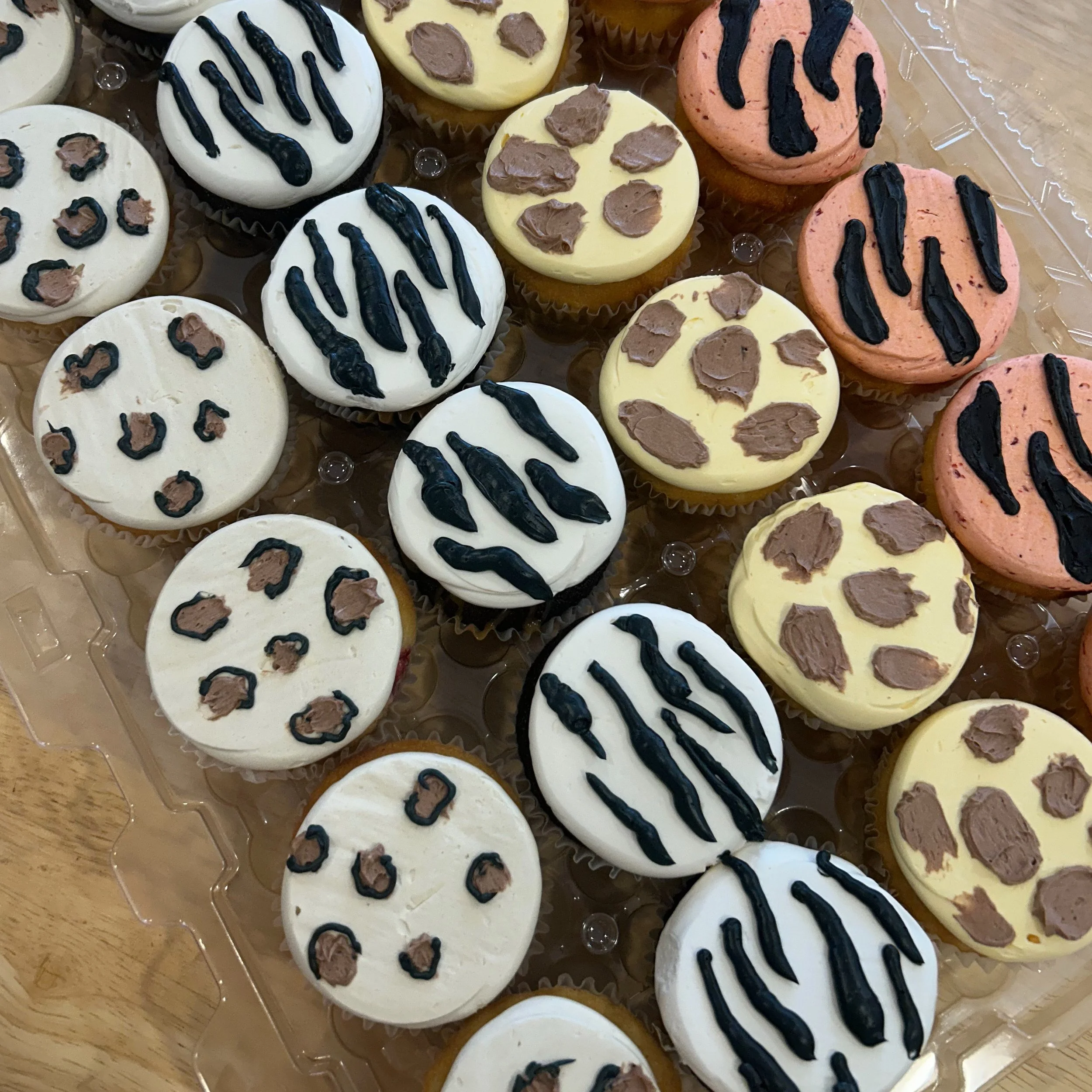 A tray of assorted cupcakes decorated with white, yellow, and pink frosting, each topped with chocolate drizzle or chocolate chips.