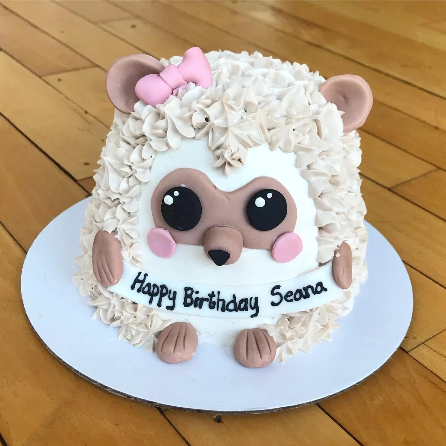 A cute fox-themed birthday cake decorated with white frosting, chocolate and pink accents, featuring a fox's face with large eyes and pink cheeks, surrounded by cream swirls and a pink bow on its head, with the message "Happy Birthday Seana" written