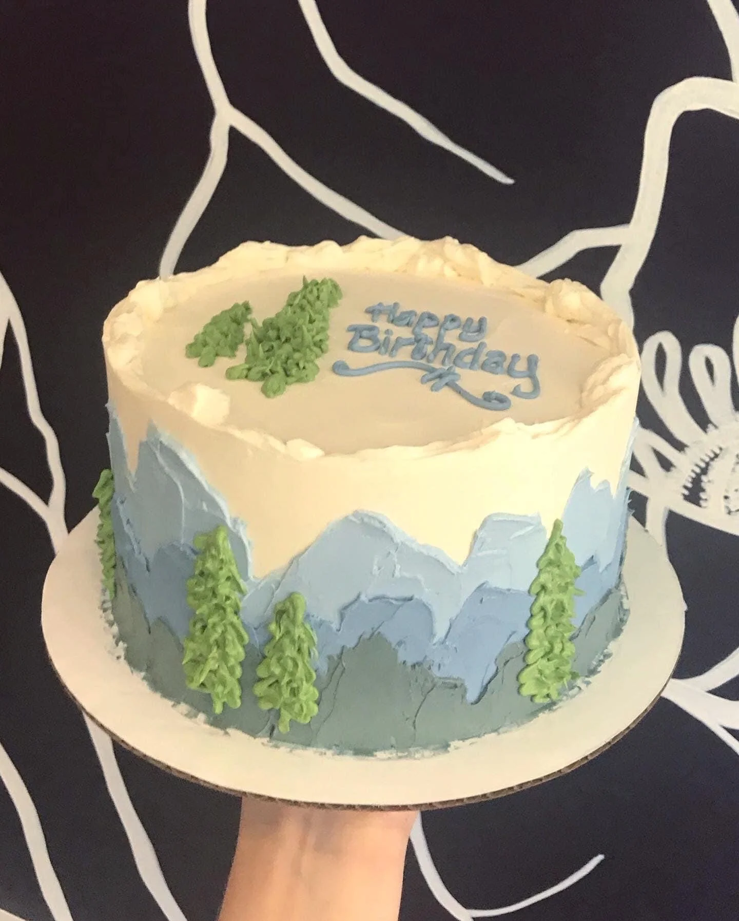 A round birthday cake with a mountain and forest theme, decorated with green trees and blue mountain peaks, with the message 'Happy Birthday' written on top in blue icing.
