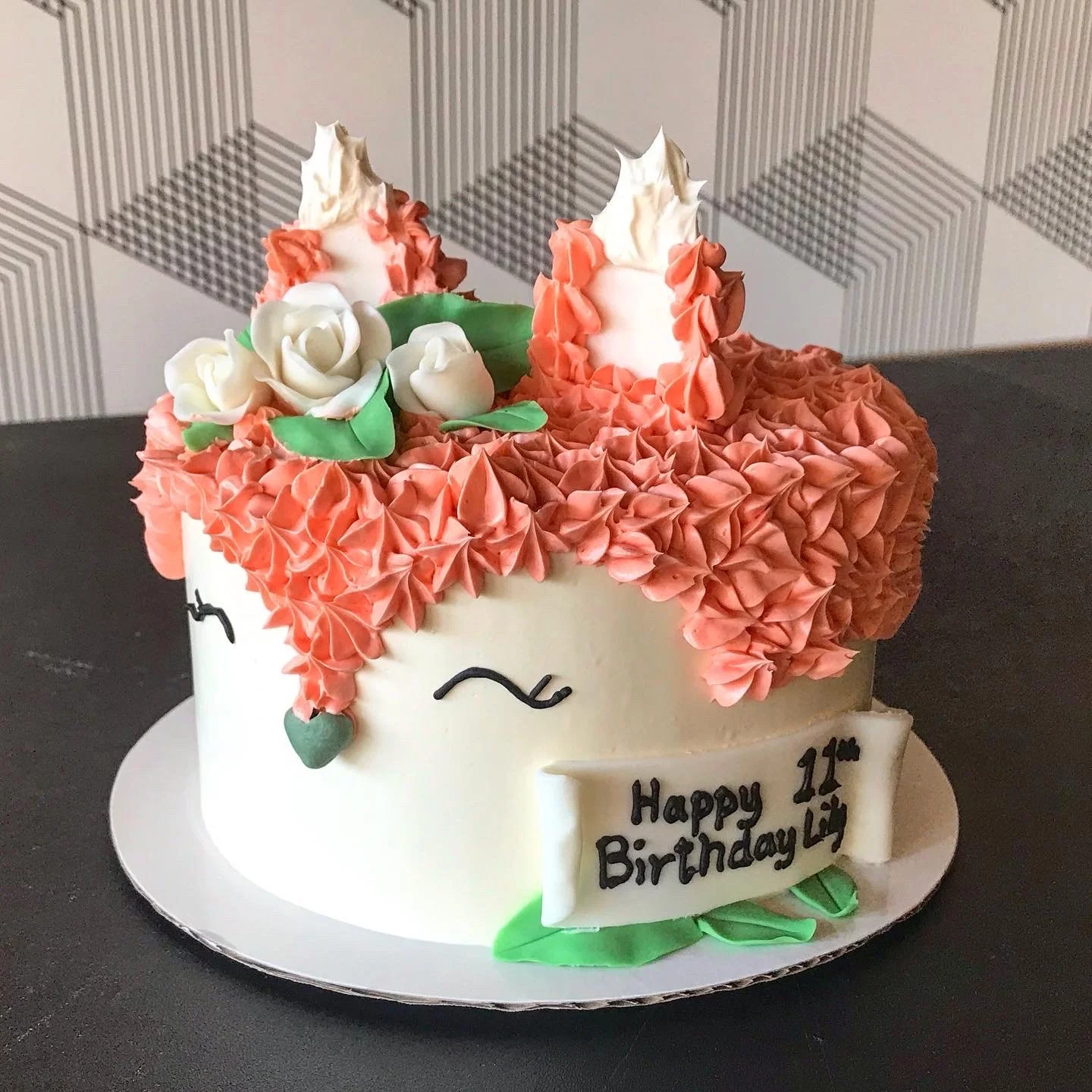 A round unicorn-themed birthday cake decorated with pink and white frosting, with closed eyes drawn on the front, a small sign that reads 'Happy 11th Birthday,' and white, pink, and green flower decorations on top.
