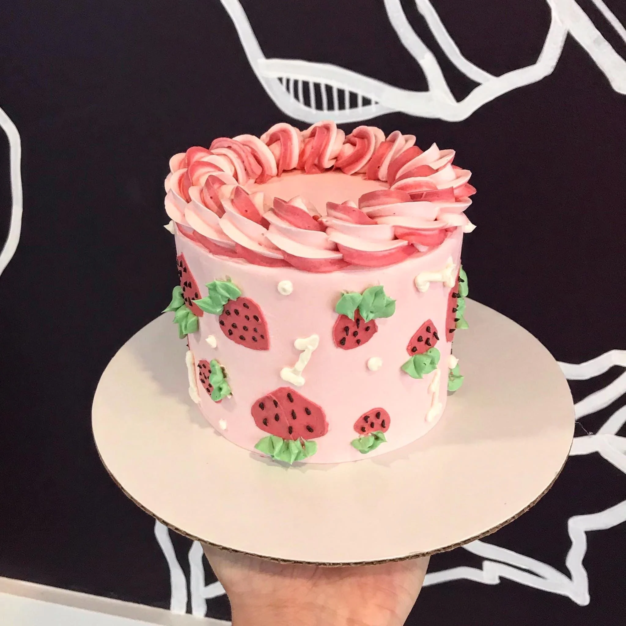 A pink birthday cake decorated with strawberry motifs, green leaves, white dots, and a pink and white piped border on top, placed on a plain round cake board.