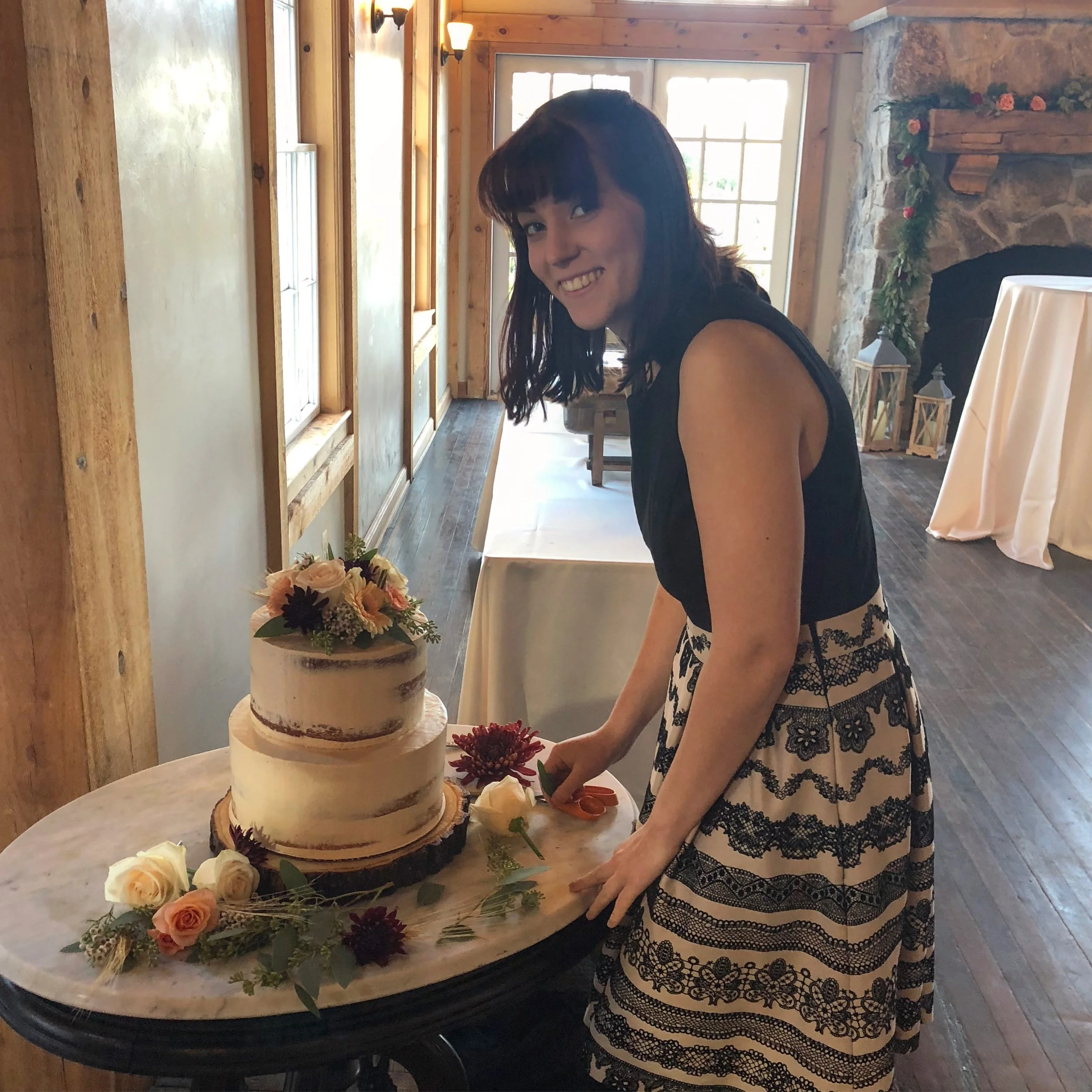 A woman with shoulder-length dark hair and bangs, wearing a sleeveless black top and a patterned skirt, is smiling while cutting a cake decorated with flowers. The cake is on a round table, and the setting appears to be a rustic indoor space with woo