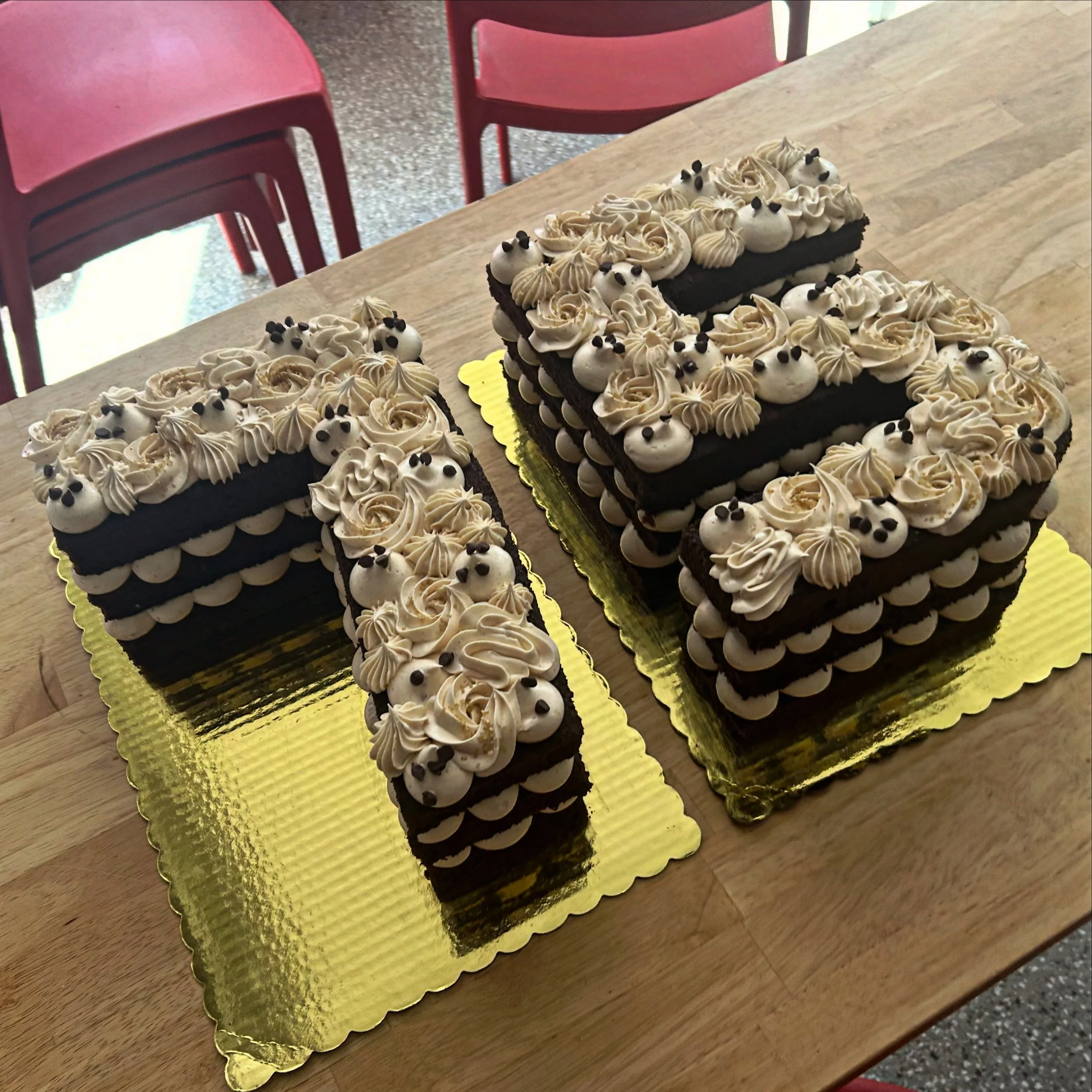 Two chocolate layered cakes decorated with white frosting and small black and white decorative accents, placed on gold-colored cake boards on a wooden table.