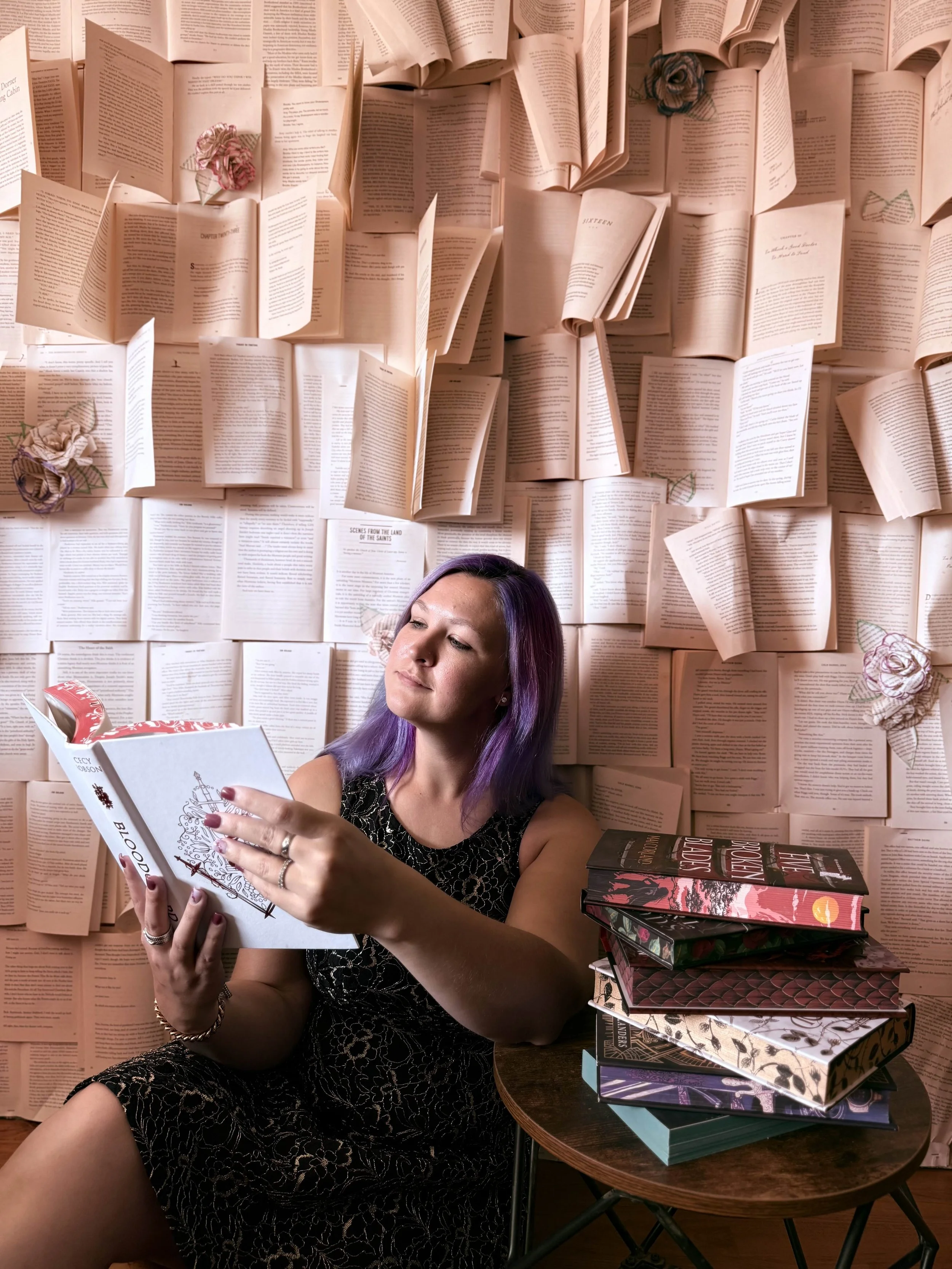 A woman with purple hair sitting against a wall covered in open books and paper flowers, reading a book, with a stack of books on a small table beside her.