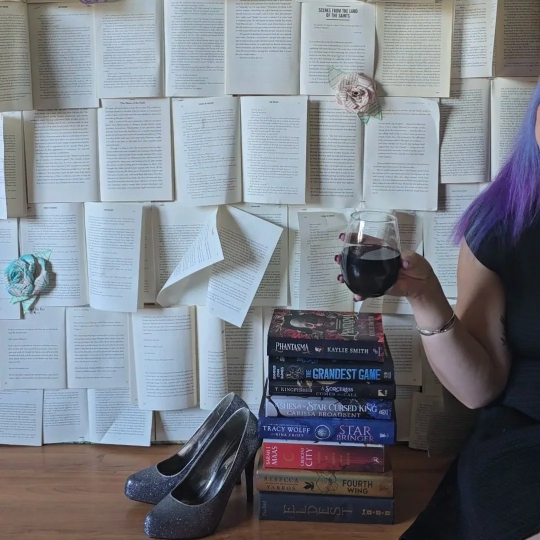 A woman with purple hair holding a glass of red wine next to a stack of books, silver high heels, and a wall decorated with open books and pages.