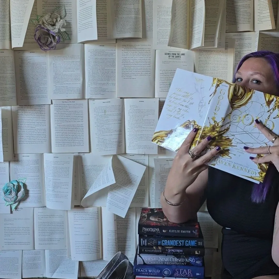 A woman with purple hair holding an open book in front of a wall covered with open books and pages. There are several stacked books on a table below her.