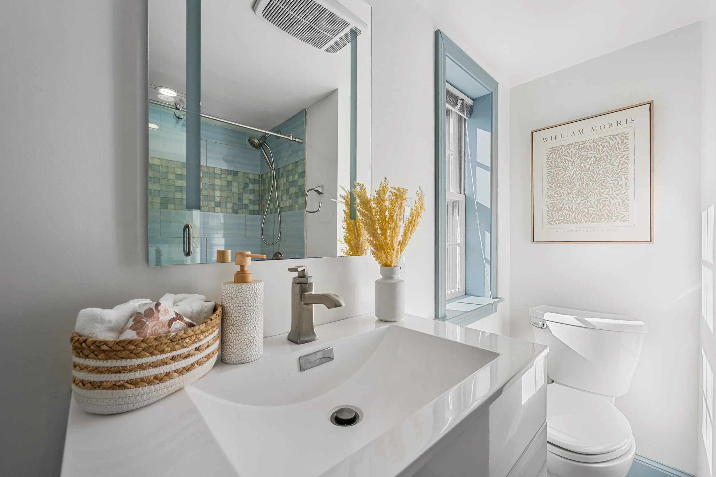 Bright bathroom with white walls, a white sink, a basket of towels, a soap dispenser, a vase with yellow flowers, a mirror, a window with blue trim, a framed artwork, and a toilet, with a view of a shower in the mirror's reflection.