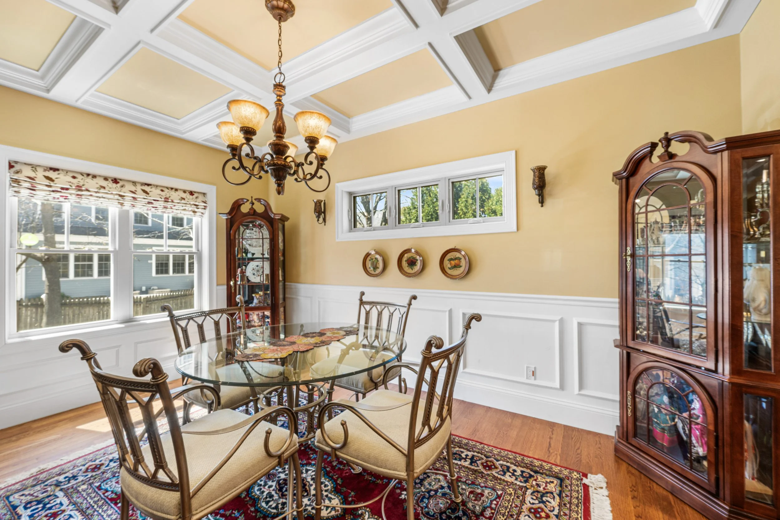 Dining room with yellow walls, white wainscoting, a chandelier, a glass-top table with six chairs, a corner cabinet, and a window with floral Roman shade.