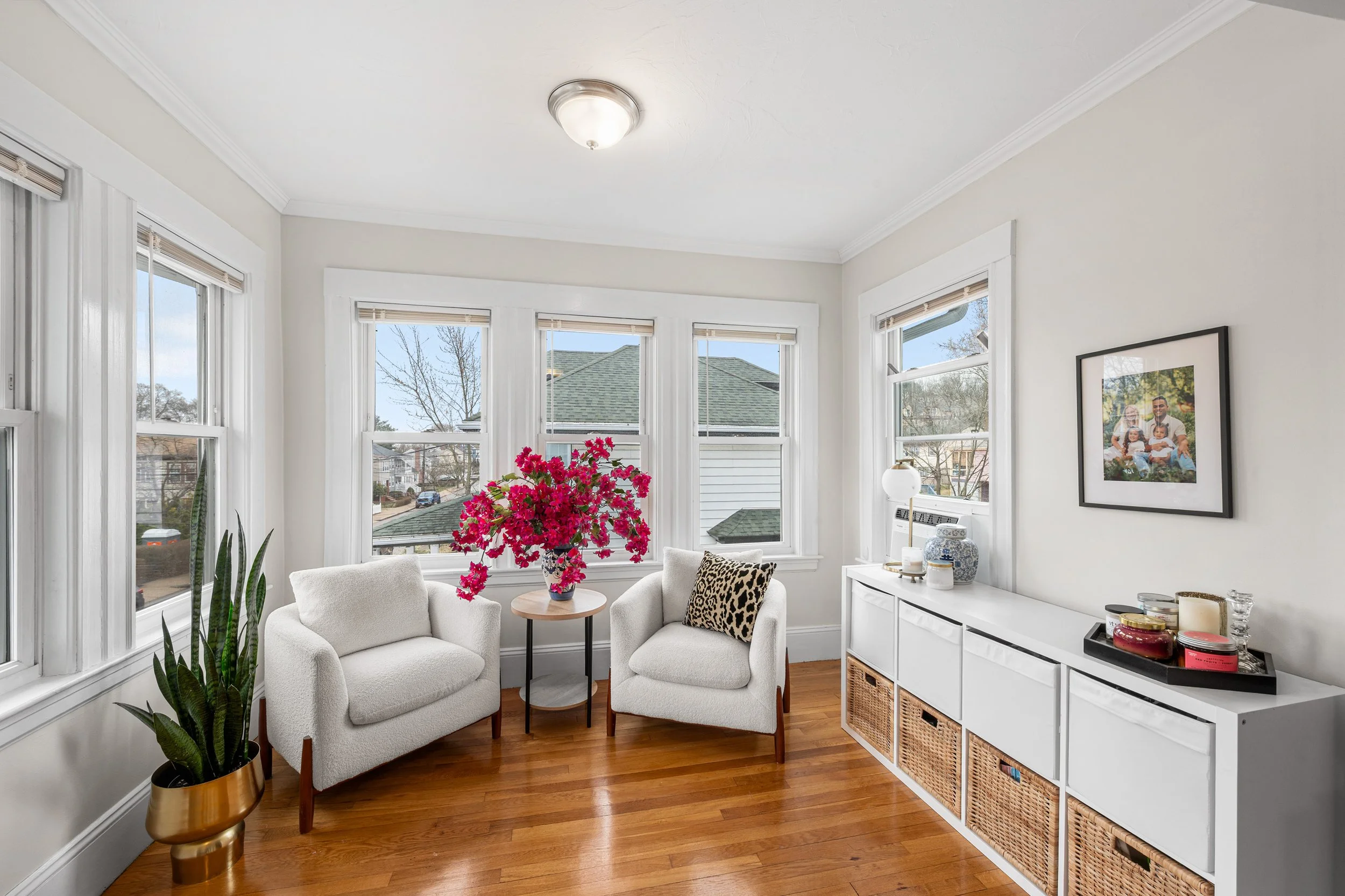 Bright sunlit living room with white walls, hardwood floors, two white armchairs, a round wooden side table with pink flowers, a potted snake plant, a white storage unit with wicker baskets, and a framed family photo on the wall.