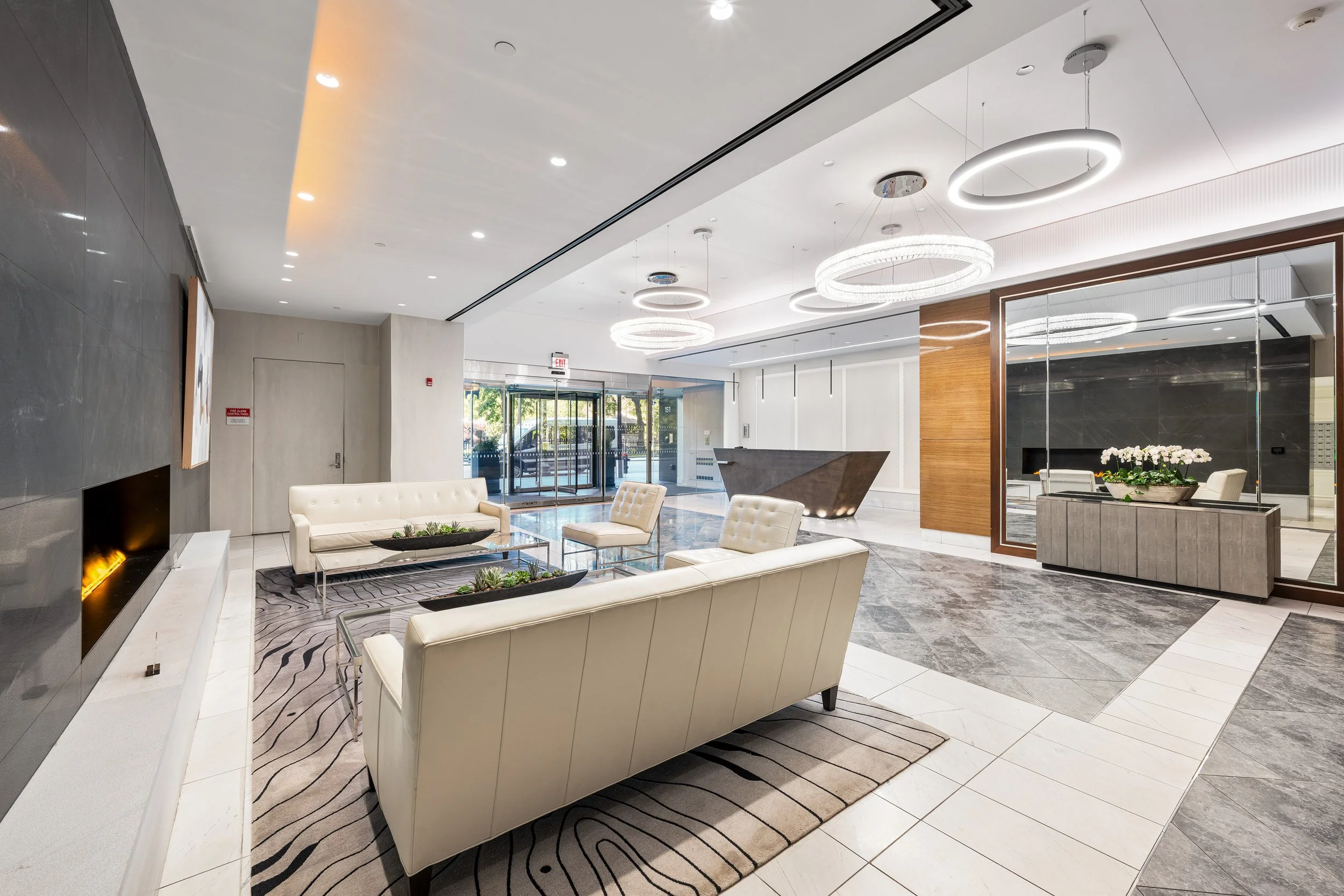 Modern hotel lobby with white sofas, glass coffee tables, a fireplace, a reception desk, and contemporary circular ceiling lights.