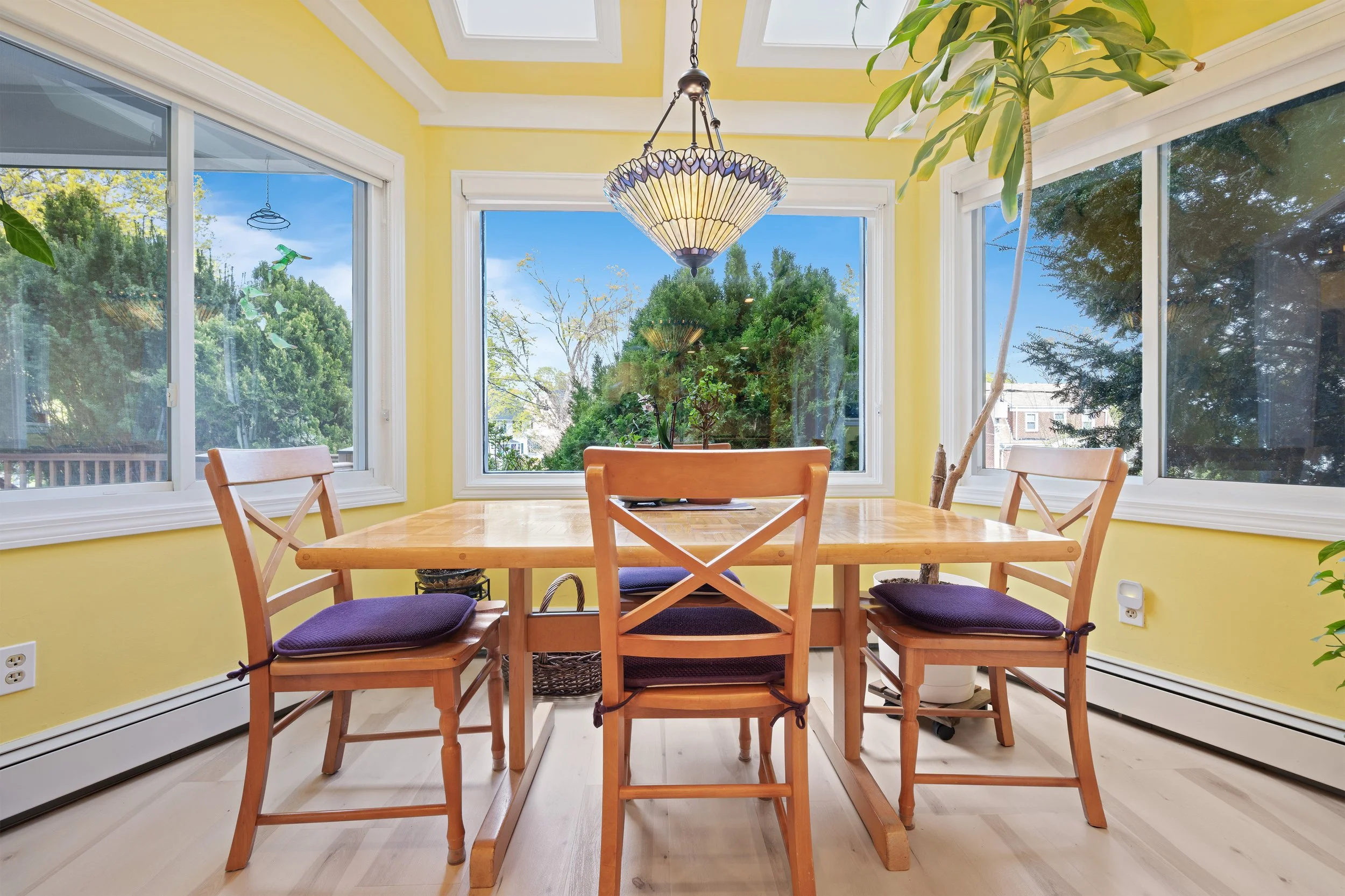 A dining area with a wooden table surrounded by four wooden chairs with purple cushions, set in a bright room with yellow walls and large windows showing trees outside. There's a woven basket under the table, a chandelier hanging from the ceiling, an