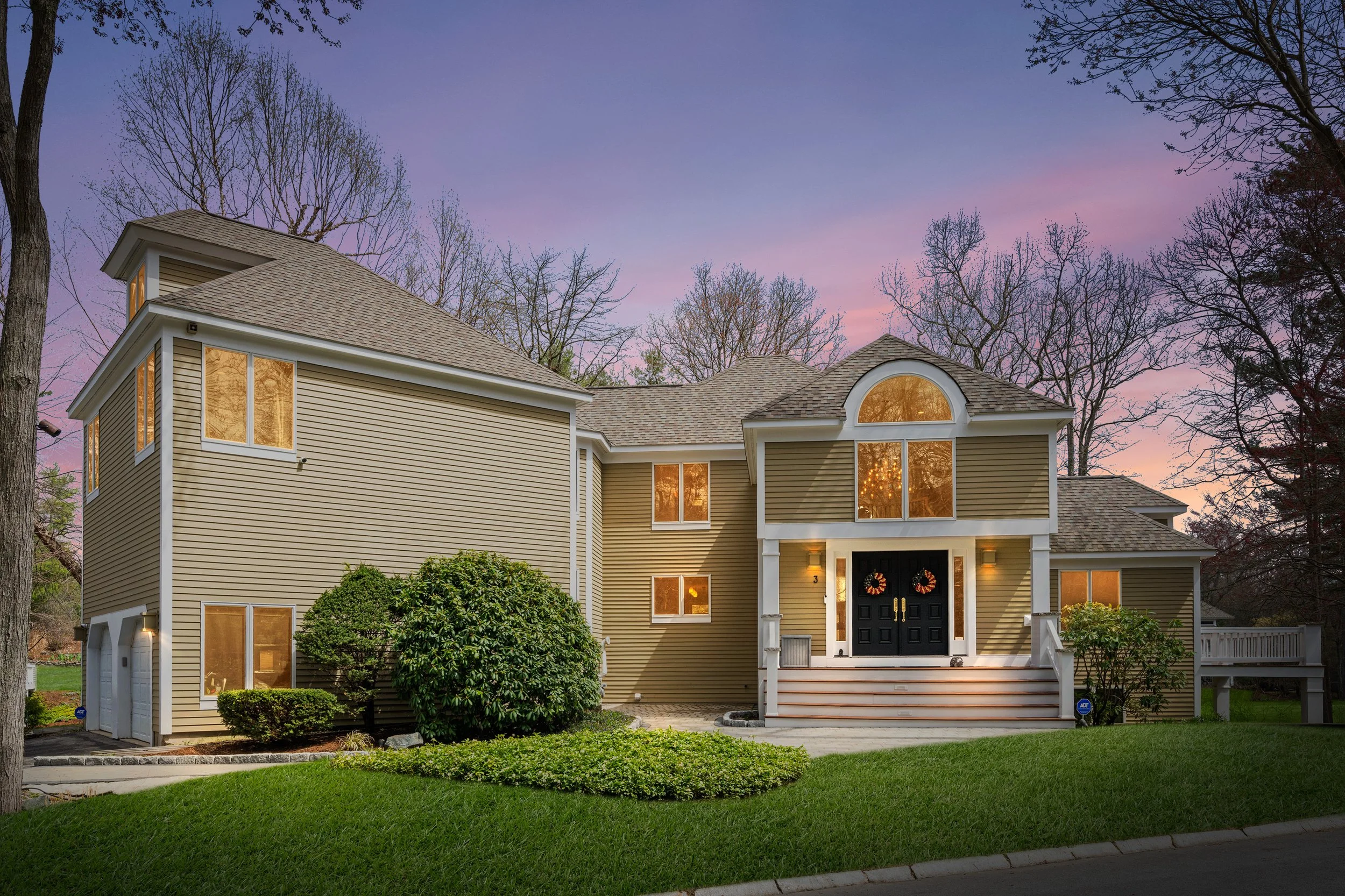 A modern two-story house with beige siding, black front door, and large arched window, illuminated by outdoor lights, surrounded by a well-maintained lawn and trees at dusk.