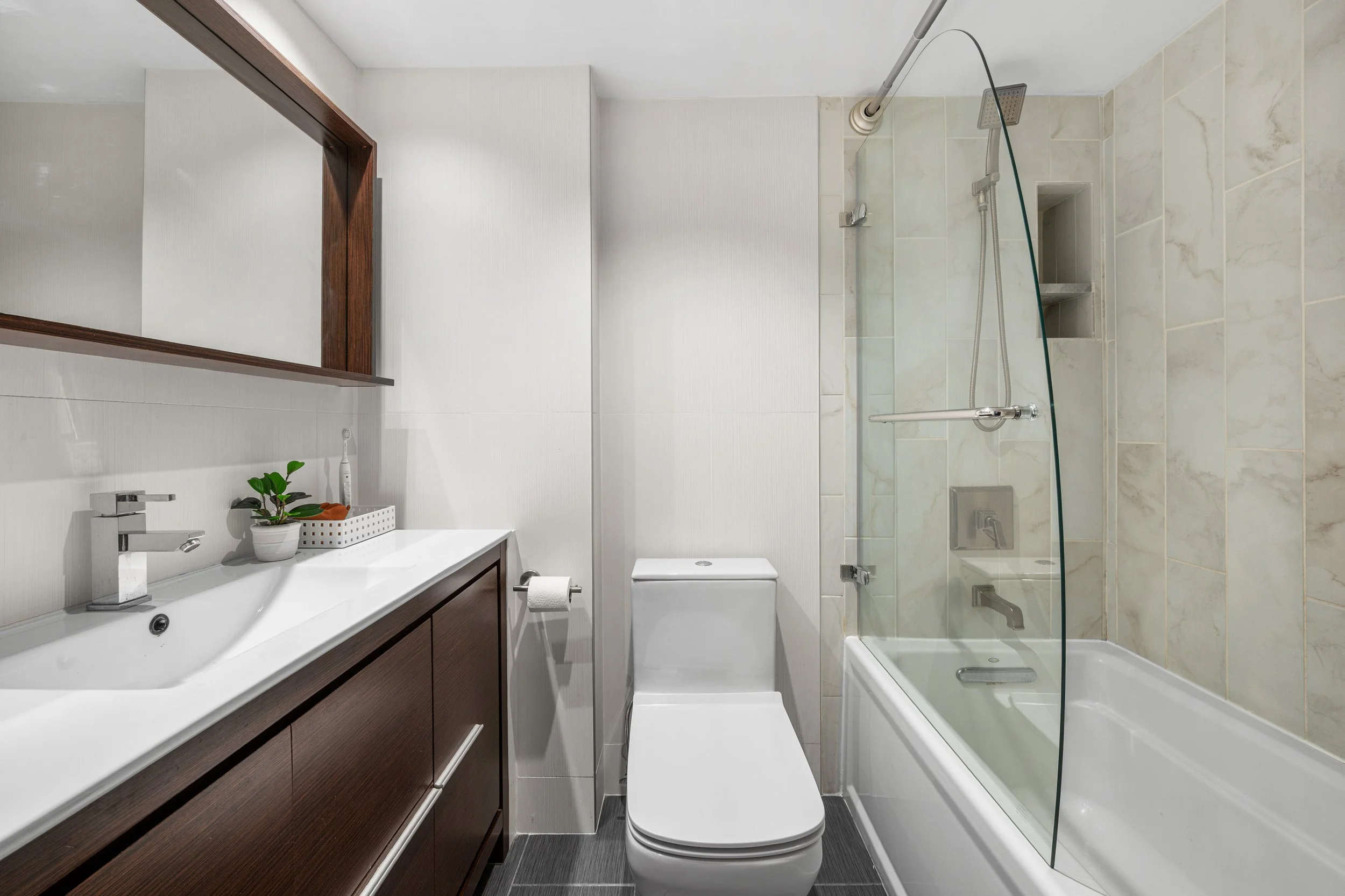 Modern small bathroom with white sink vanity, bathroom mirror, green potted plant, toilet, and a shower with glass door and beige tiles.