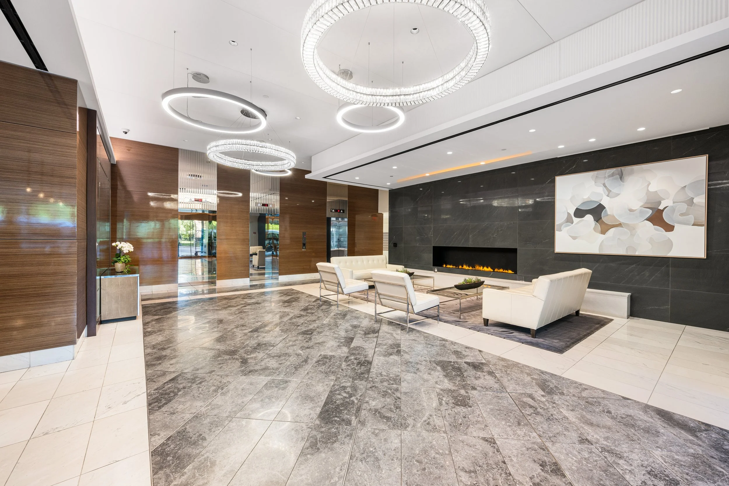 Modern hotel lobby with marble flooring, white seating, a wall-mounted fireplace, abstract artwork, and contemporary circular chandeliers.