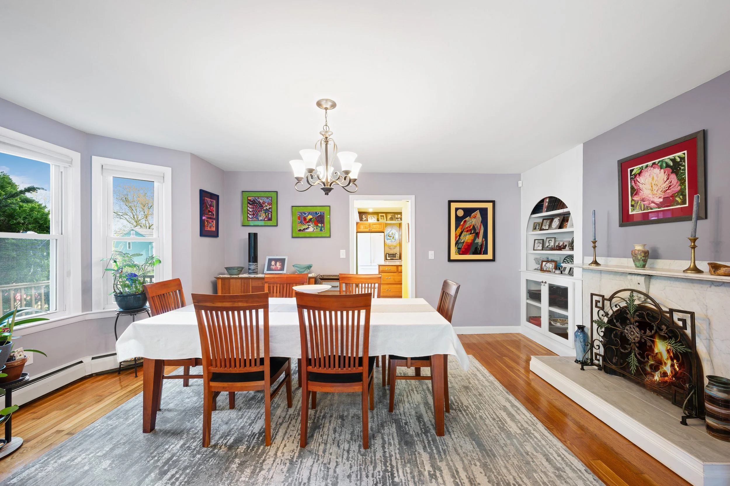 Dining room with wood floor, purple walls, a white marble fireplace with a decorative screen, a chandelier, six wooden chairs around a white table, colorful framed artwork on the walls, large windows with plants, and a doorway leading to a bright kit