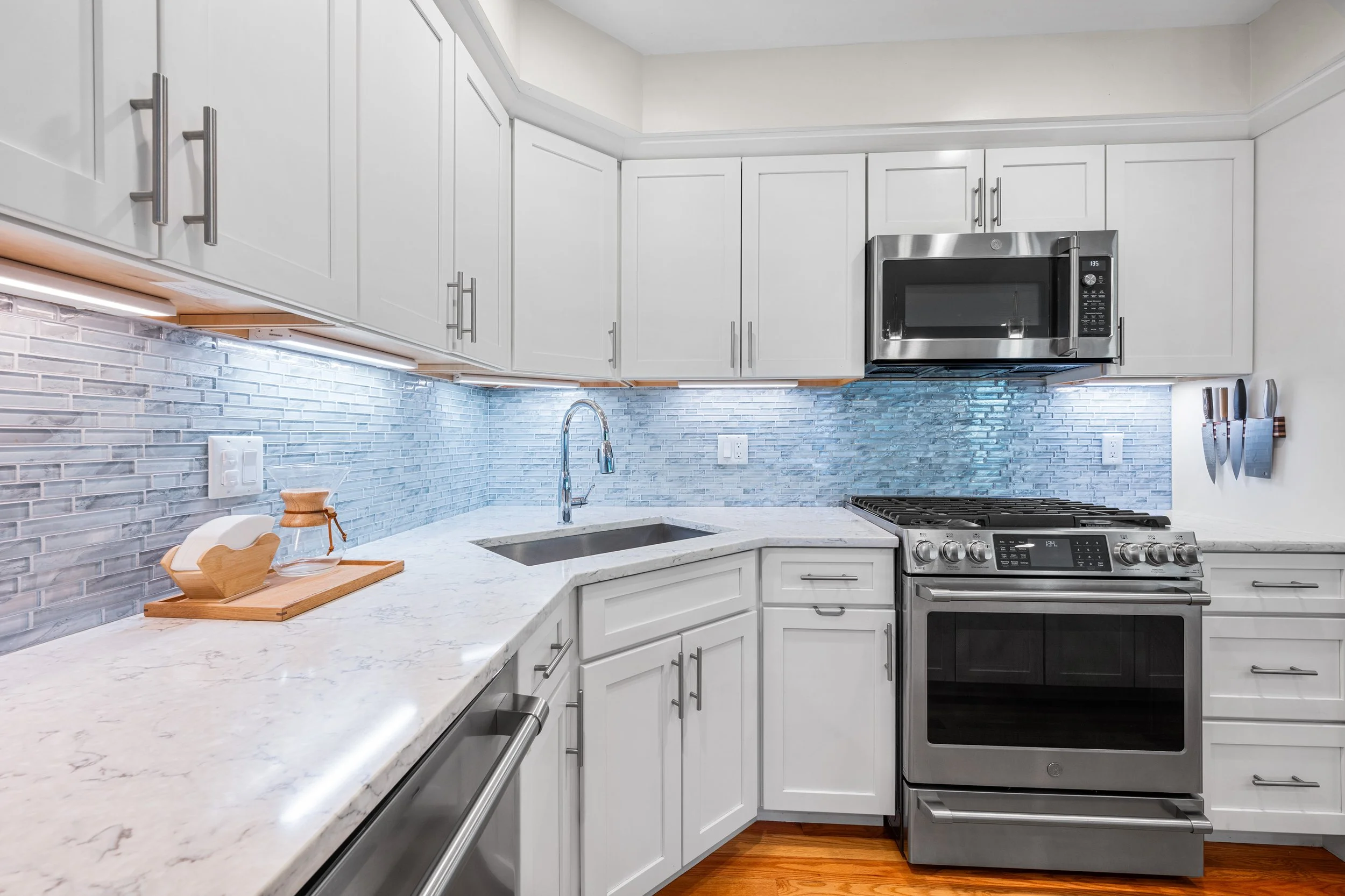 White kitchen with marble countertops, gray backsplash, stainless steel appliances including microwave, oven, and dishwasher, with a small knife rack on the wall.