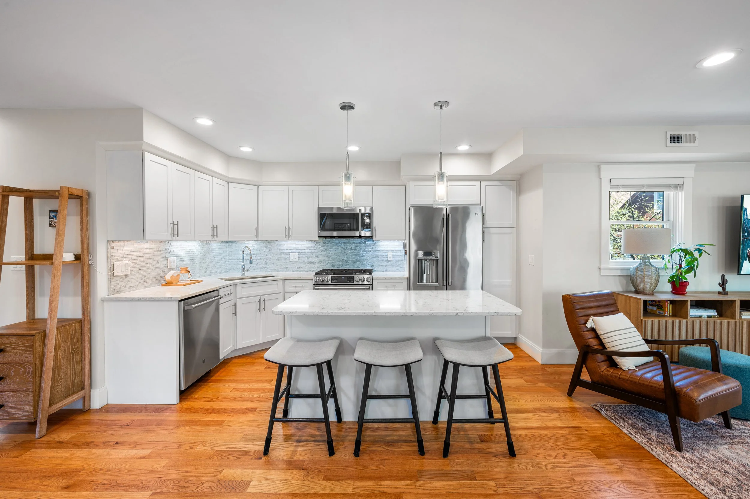 Modern kitchen with white cabinets, marble island, stainless steel appliances, hardwood floors, and adjacent living space with armchair and side table.