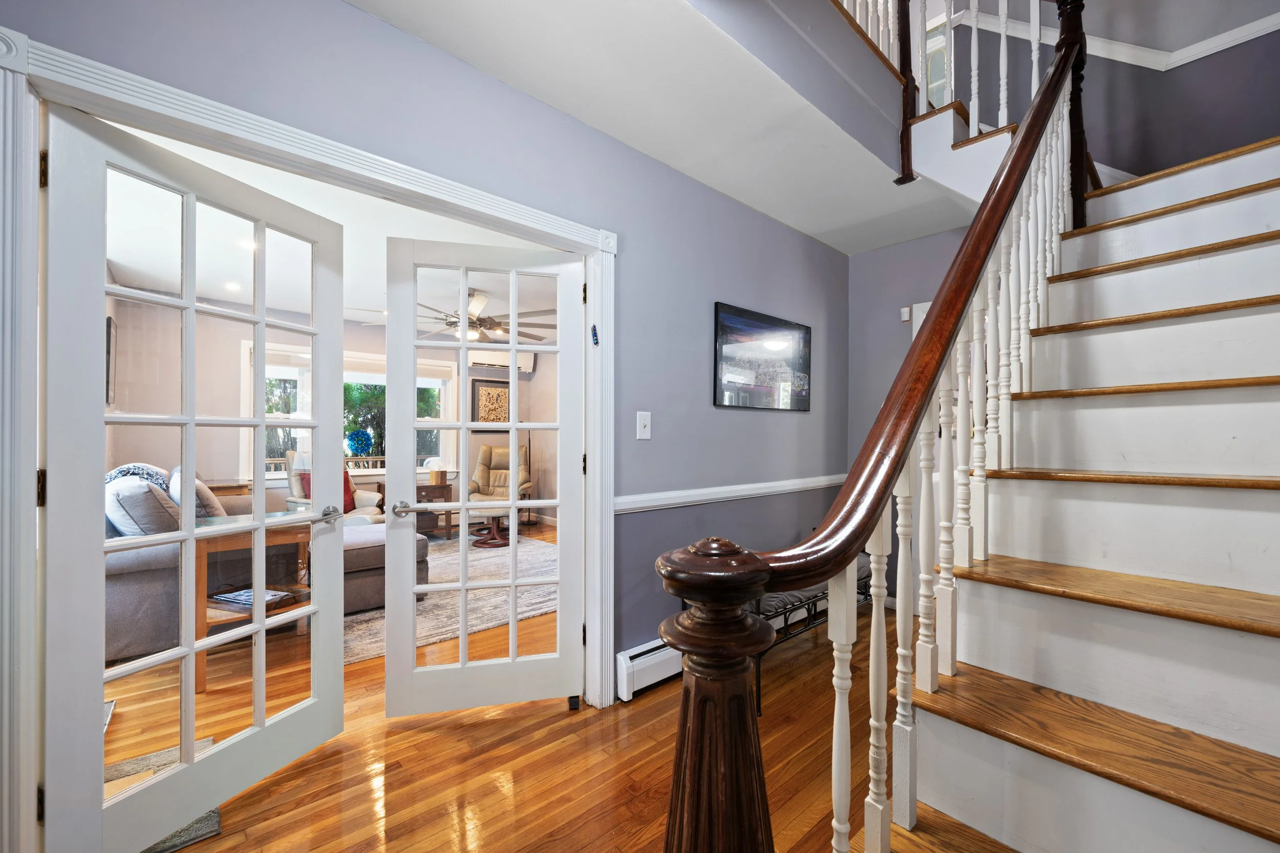 Interior view of a house with a staircase, hardwood floor, and a living room visible through open French doors.