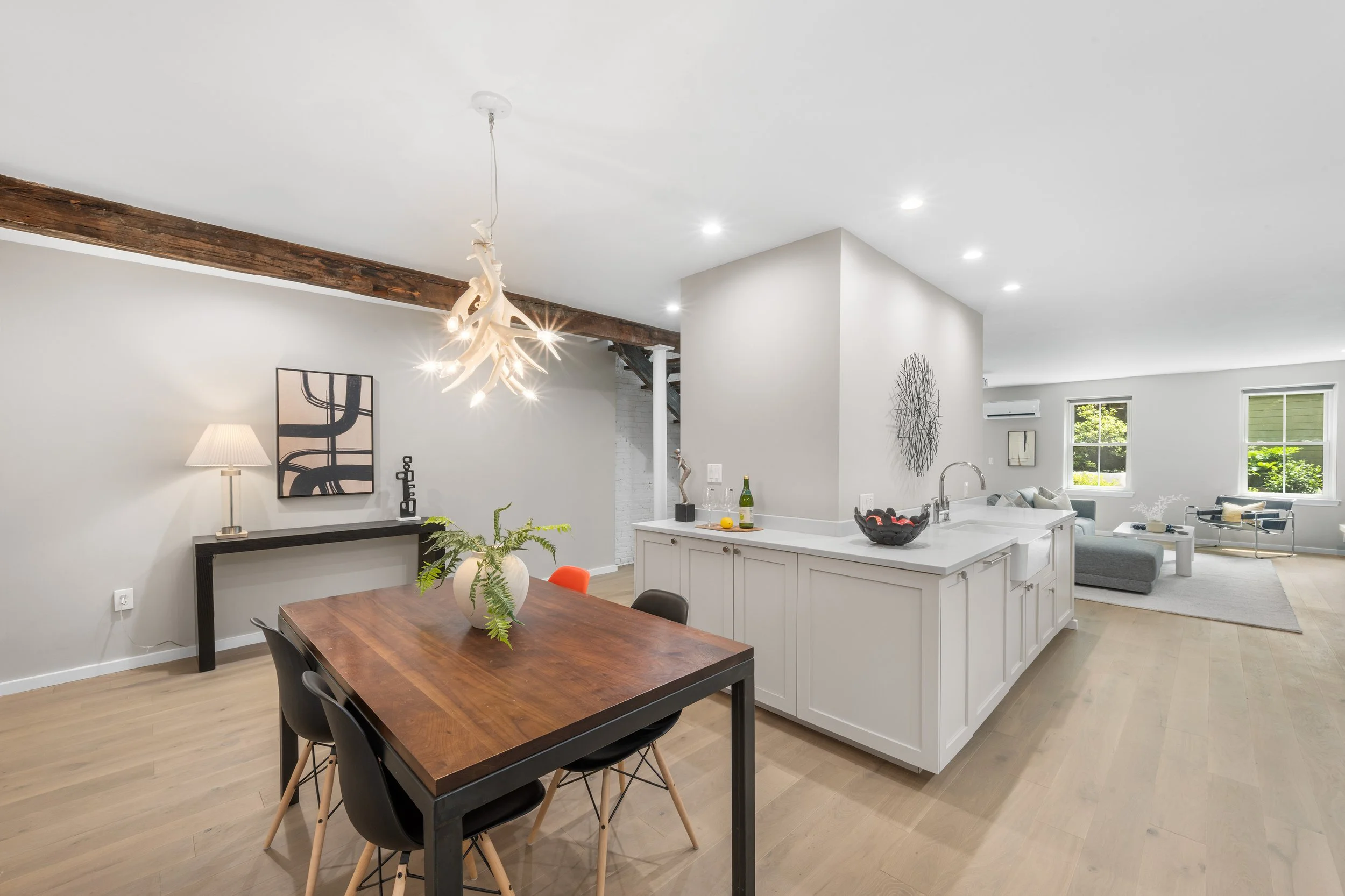 Open-concept living and dining area with a wooden dining table, black chairs, white kitchen island, modern decor, and large windows showing green outdoor scenery.