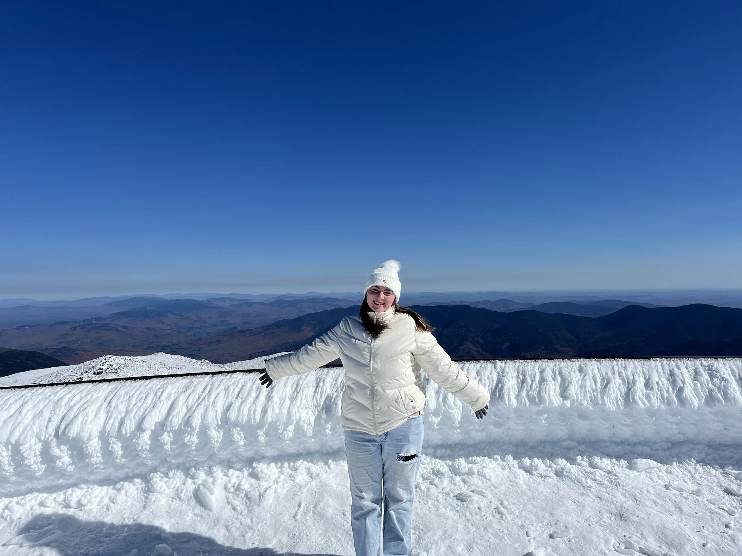A woman smiling and dressed in winter clothing standing on snow atop a mountain with panoramic view of distant mountain ranges and a clear blue sky.