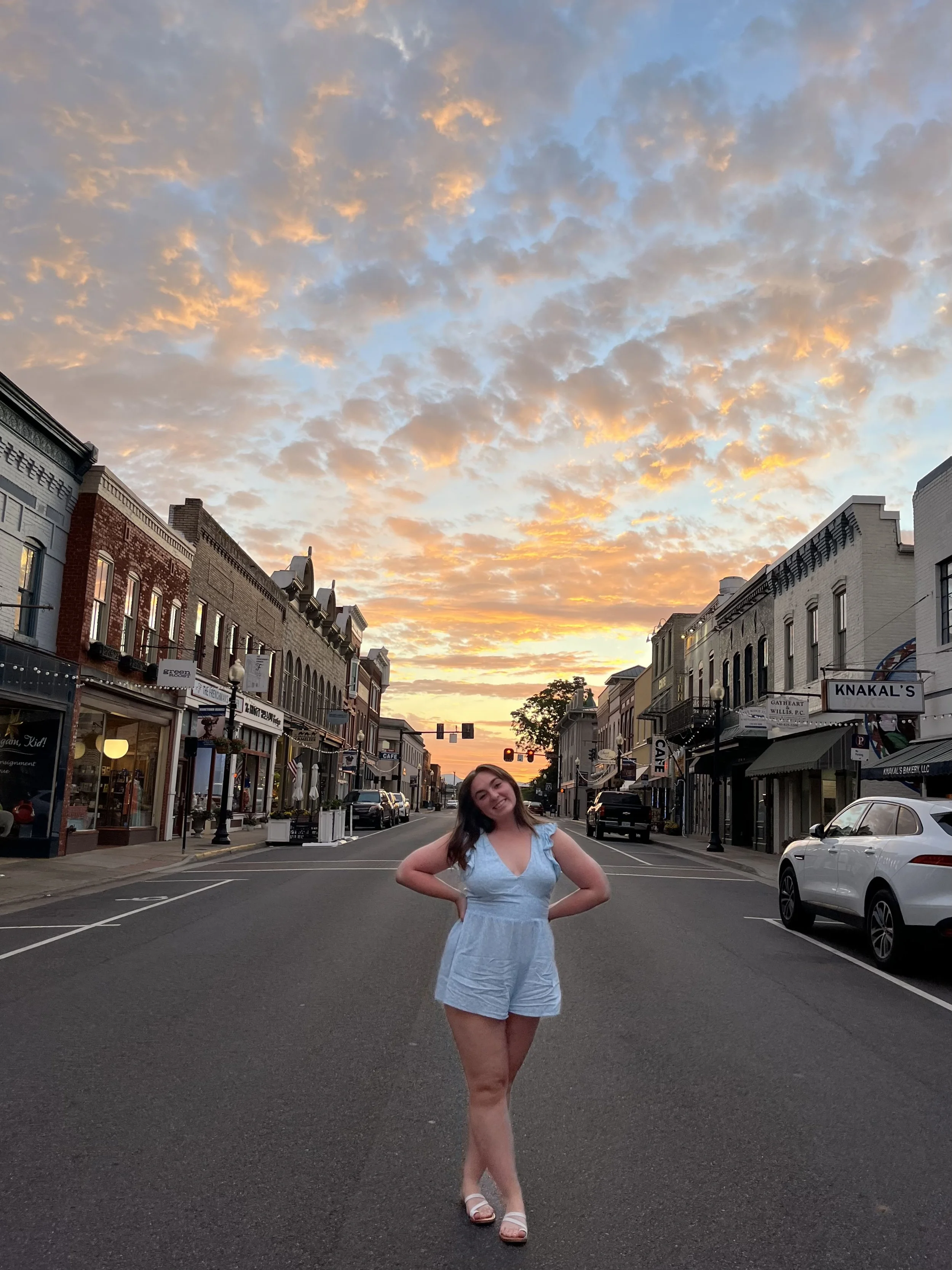 A woman in a light blue romper stands in the middle of a city street during sunset, with a sky filled with orange and pink clouds behind her. The street is lined with small shops, some with signs and awnings, and a few parked cars.