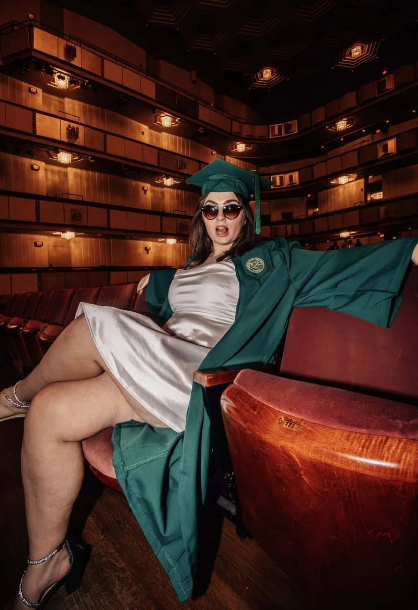 A woman in a graduation cap and gown sitting alone in an auditorium with red theater seats and wood-paneled walls, wearing sunglasses and a satin dress.