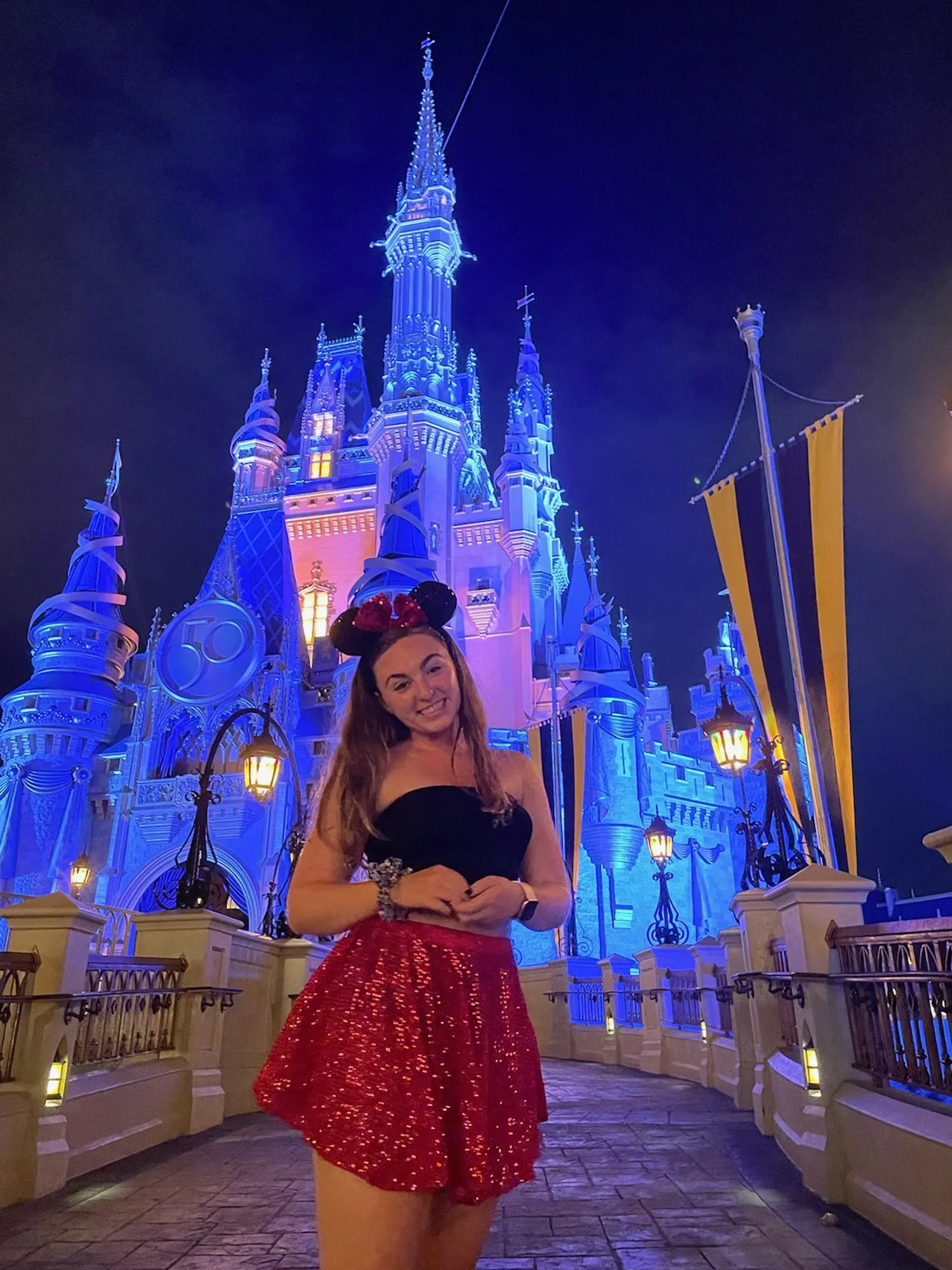 A young woman wearing black top, red sequined skirt, and Minnie Mouse ears hat standing in front of a brightly lit castle at night.