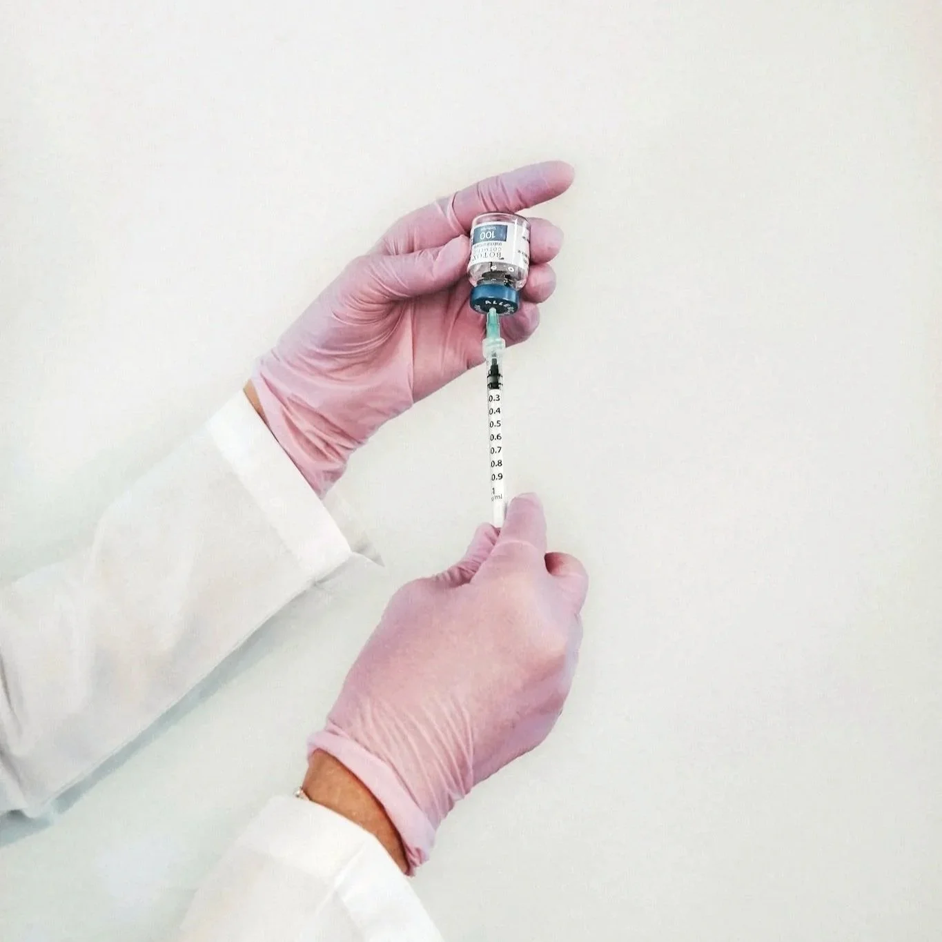 A healthcare worker wearing pink gloves and a white lab coat drawing a vaccine from a vial into a syringe.