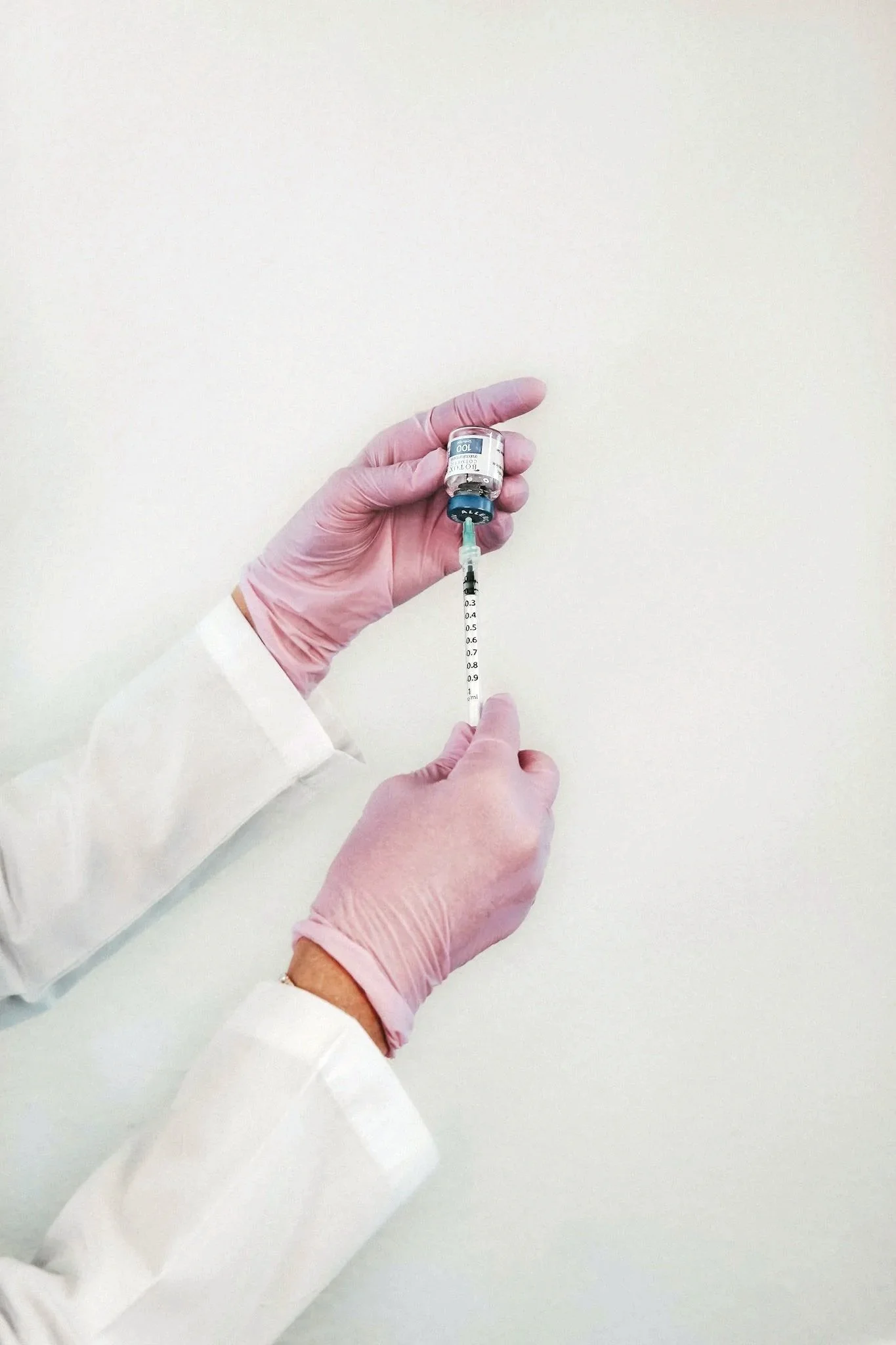 A person wearing pink gloves and a white lab coat holding a botox vial and drawing botox into a syringe.