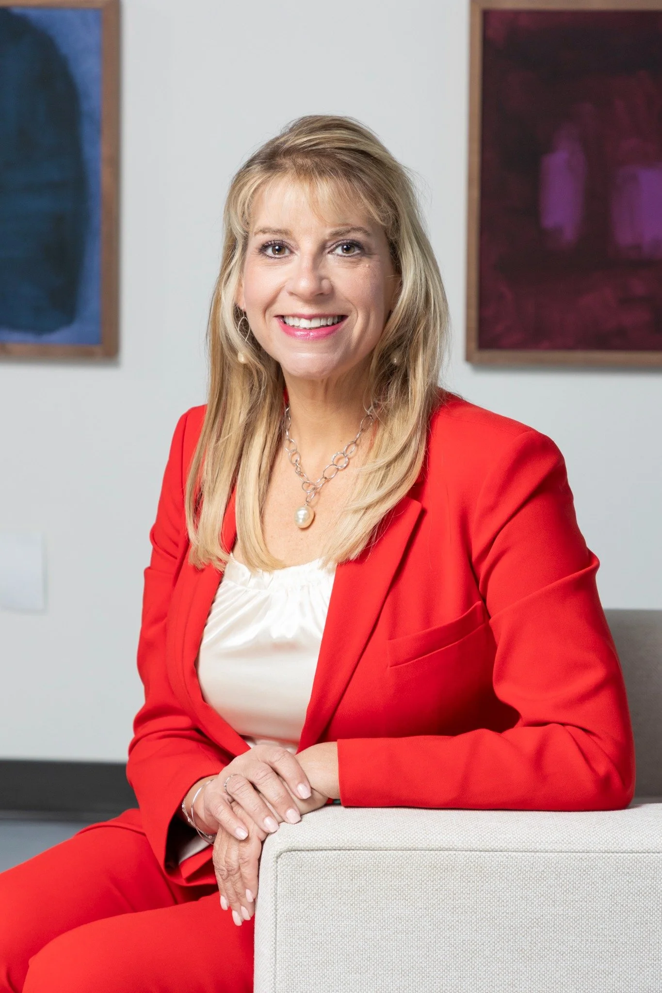 A woman with blonde hair smiling, wearing a red blazer and white top, sitting on a light-colored seat, with colorful abstract artwork on the wall behind her.