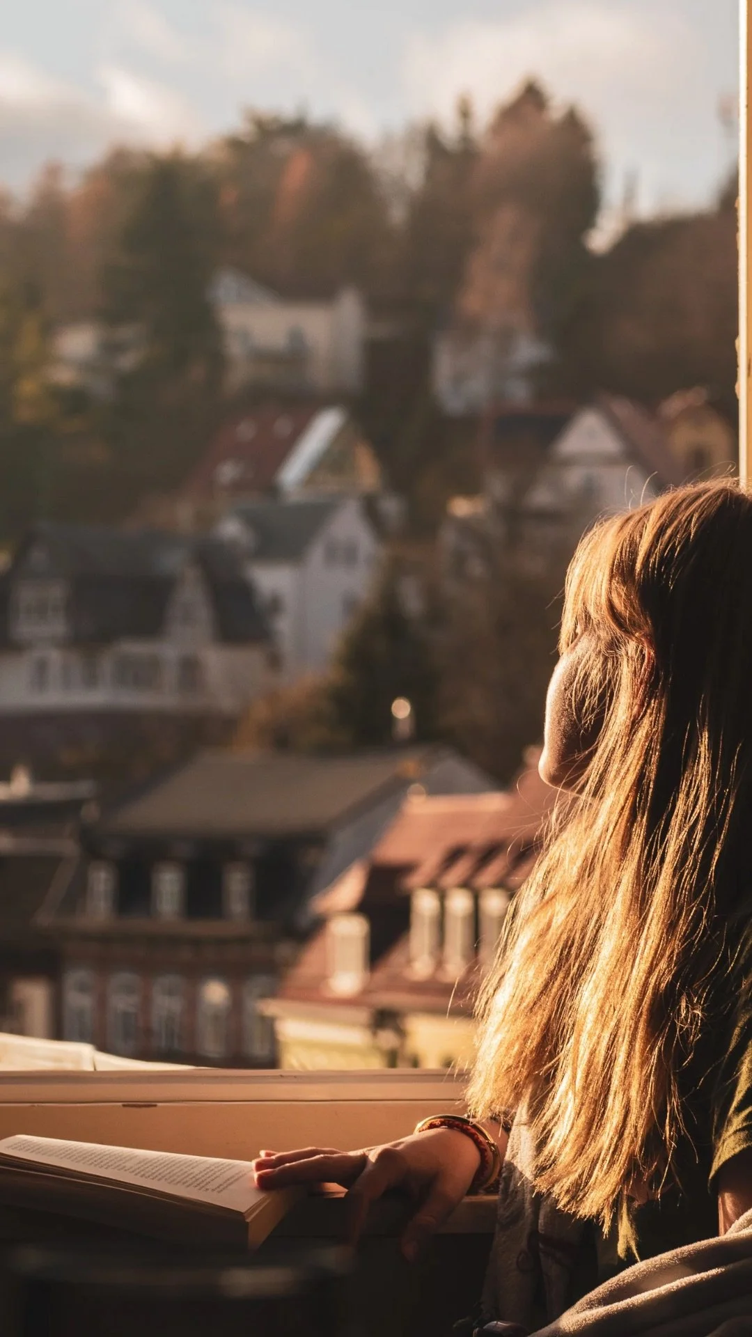 A girl with long light brown hair reading a book near a window in sunlight, with a view of houses and trees outside.
