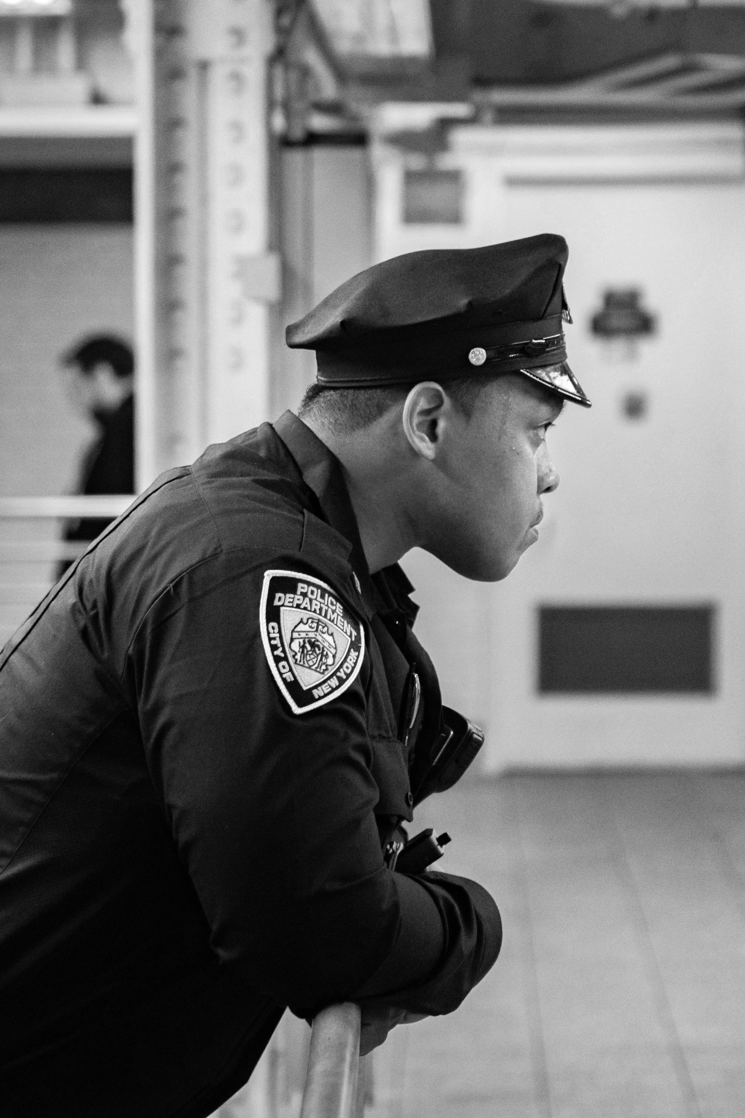A young police officer wearing a uniform and peaked cap, leaning on a railing in an indoor setting.
